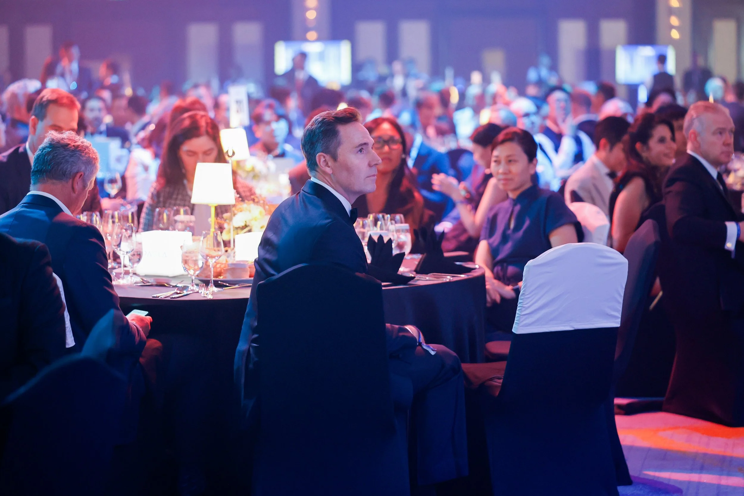 A group of people dressed in formal attire attending a banquet or gala, seated at tables with wine glasses and floral centerpieces, under dim, colorful lighting in a large hall.