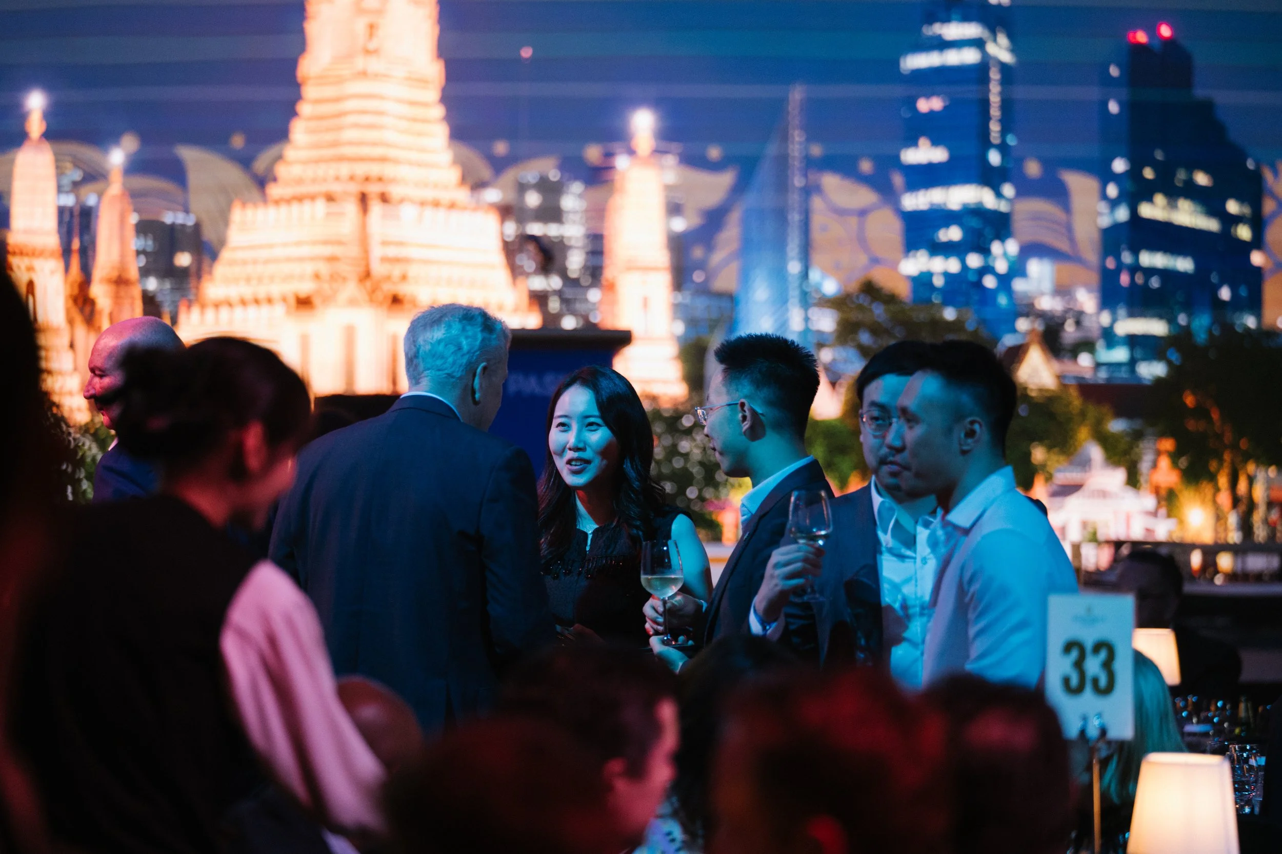 People gathered at an outdoor evening event with a city skyline featuring illuminated buildings and a tall, lit-up statue in the background.
