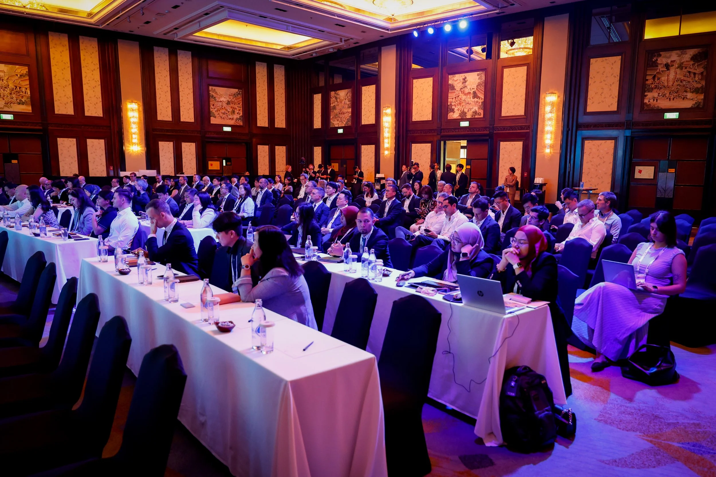 A large conference room filled with seated attendees dressed in business attire, facing forward with some taking notes or using electronic devices. The room has high wooden-paneled walls with decorative art panels, warm lighting, and a high ceiling with a large light fixture. There are tables with bottles of water and conference materials.