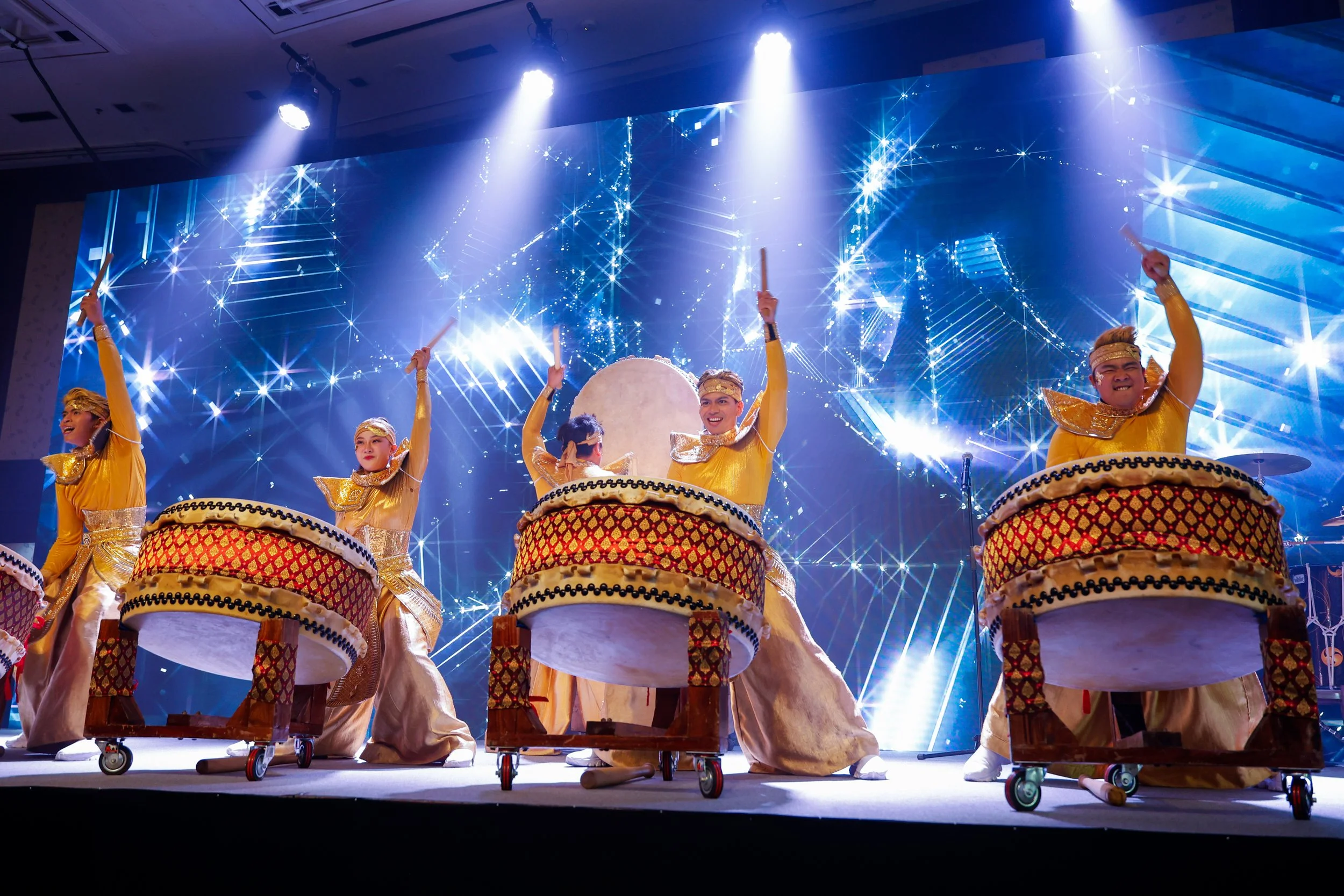 Performers dressed in gold costumes playing large traditional drums on stage with digital light effects in the background.