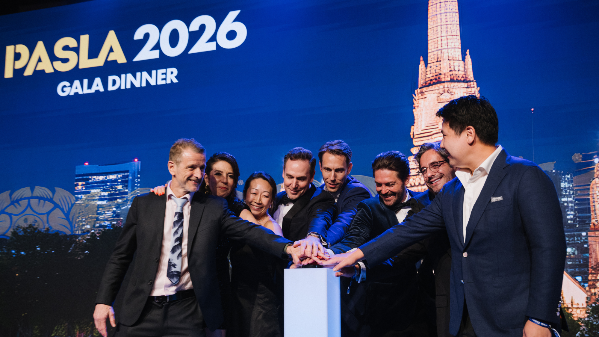 A group of people in formal attire participating in a ceremonial event, all placing their hands together over a pedestal during the PASLA 2026 Gala Dinner, with a lit-up cityscape and a prominent tower in the background.