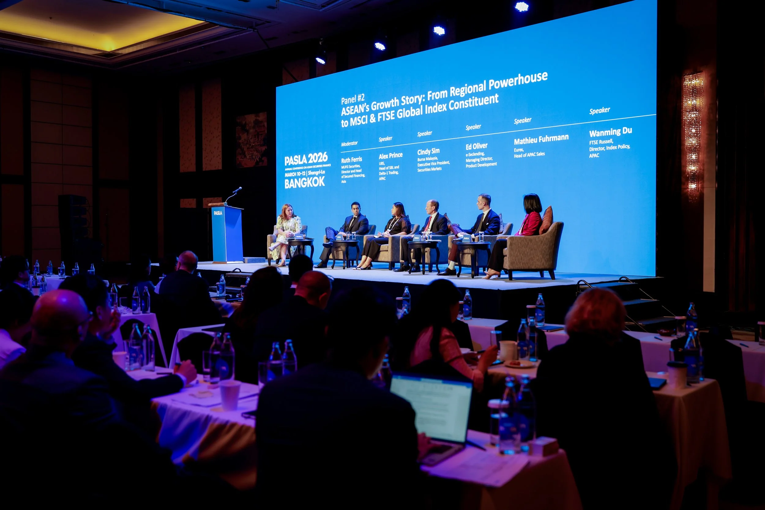 A panel discussion at the PASLA 2026 conference in Bangkok. Six speakers and a moderator are seated on stage, and a large screen behind them displays the event details and speaker names. The audience is seated at tables in front of the stage.