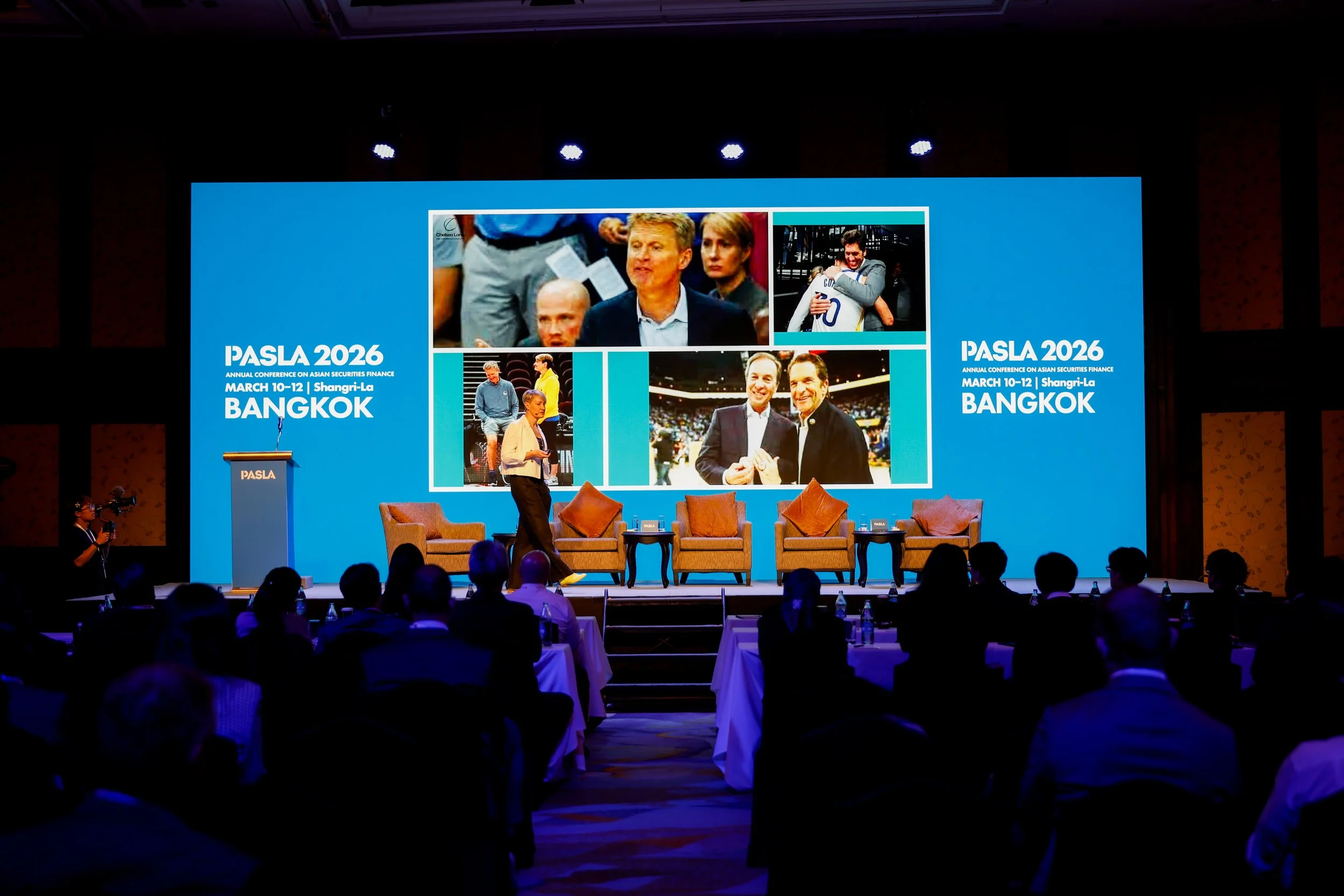 Large conference stage with a big screen displaying images of people. The screen mentions PASLA 2026, an annual conference on Asian securities finance, held from March 10-12 in Bangkok. There are chairs on the stage, and an audience seated in front.