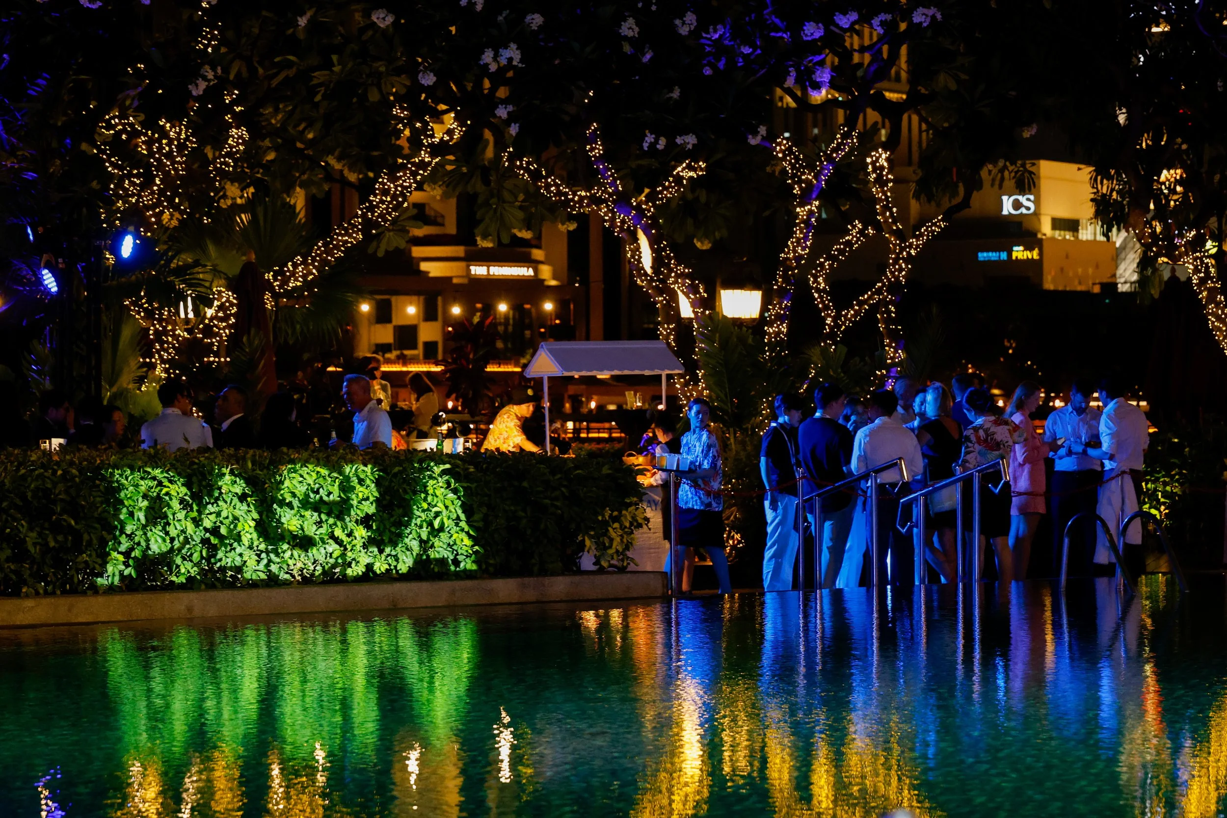 Night view of an outdoor gathering with people standing along a riverbank. The trees are decorated with string lights, reflecting colorful lights on the water. Buildings and signs are visible in the background.