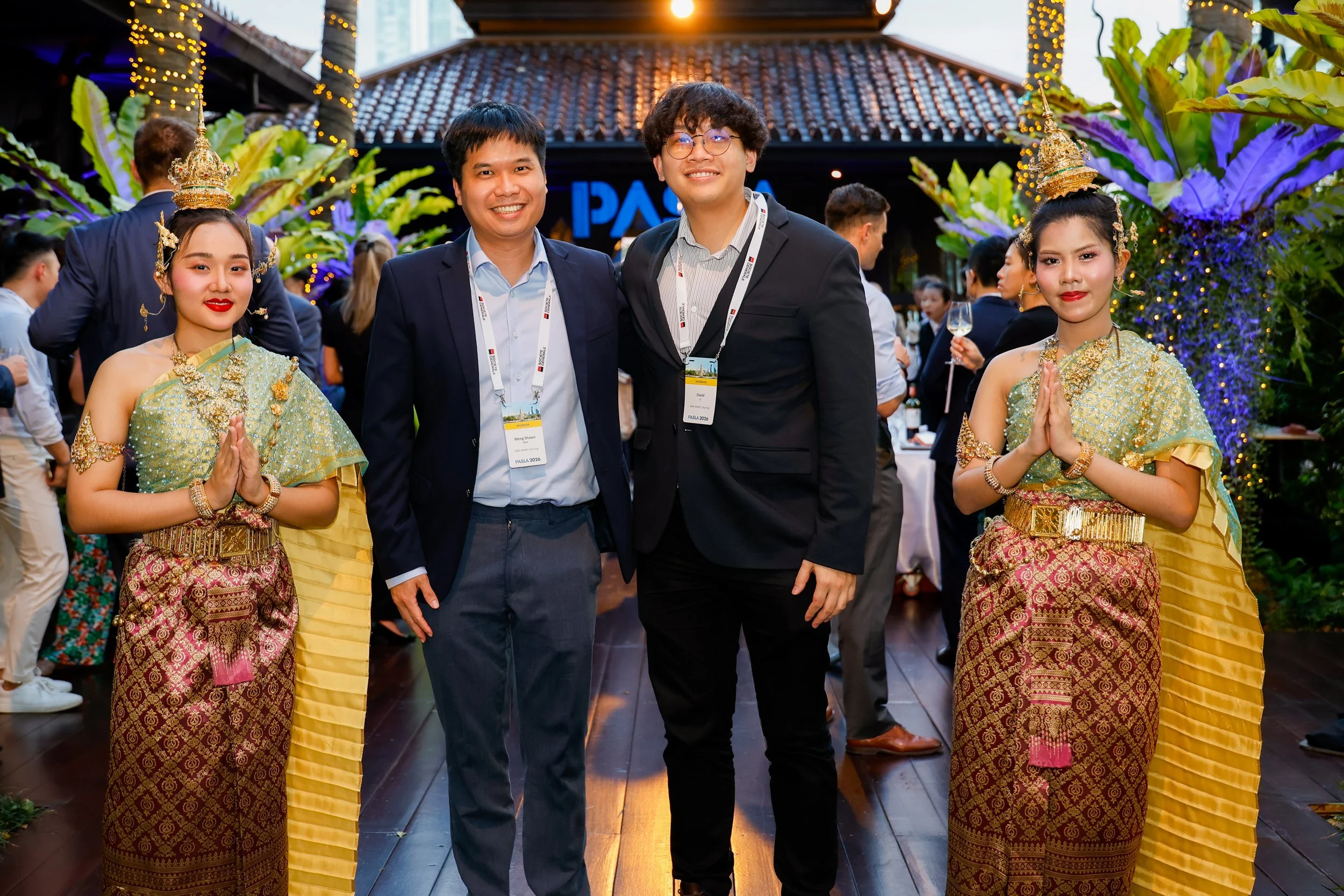 Two men in suits stand between two women dressed in traditional Thai attire with gold jewelry and decorative headdresses, at an outdoor event with tropical plants and string lights.