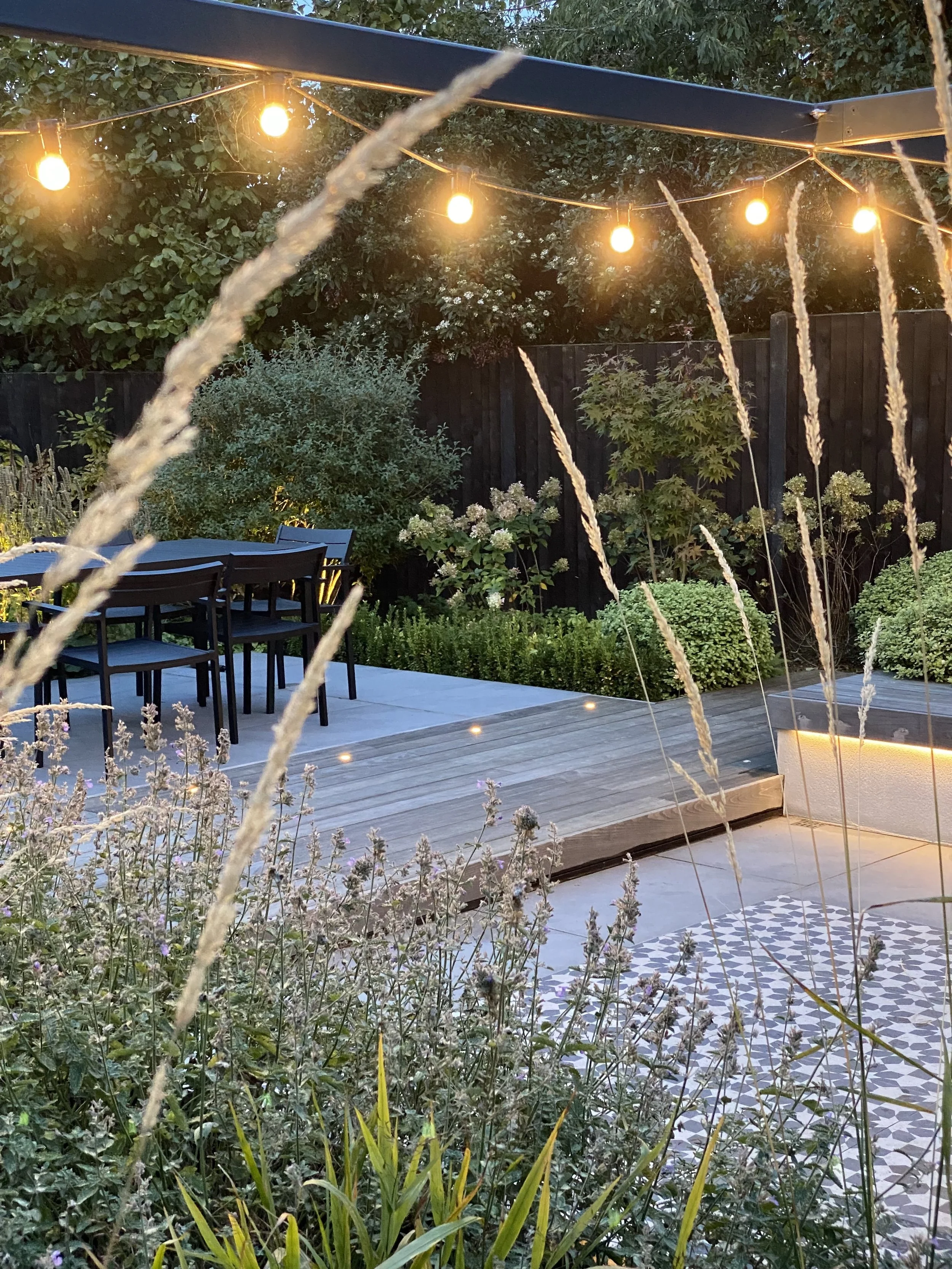 A cosy garden patio at dusk with string lights hanging above a wooden deck and outdoor dining table with chairs, surrounded by lush green plants and a black fence.