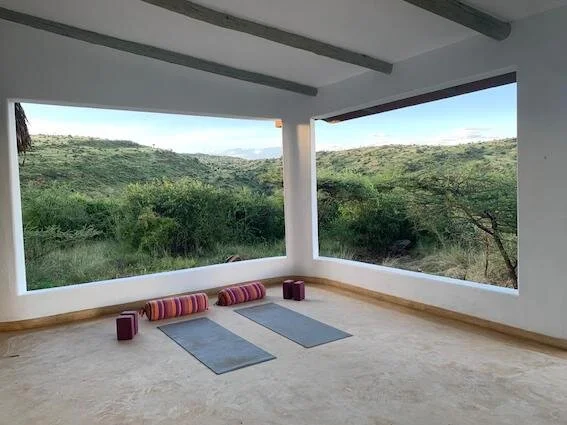 Indoor yoga or meditation space with large open windows overlooking a green hillside landscape, two yoga mats, bolsters, and blocks on a light-colored floor.