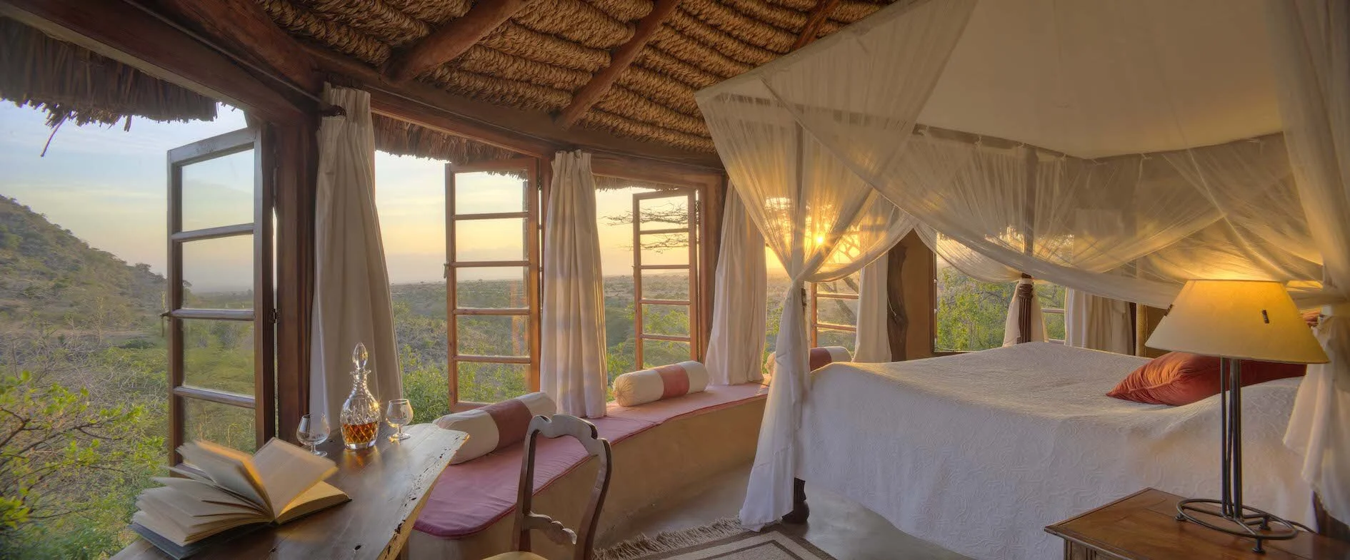 A cozy bedroom with a four-poster bed under a mosquito net, and open windows with curtains that reveal a scenic view of a sunset over a rural landscape with trees and hills.