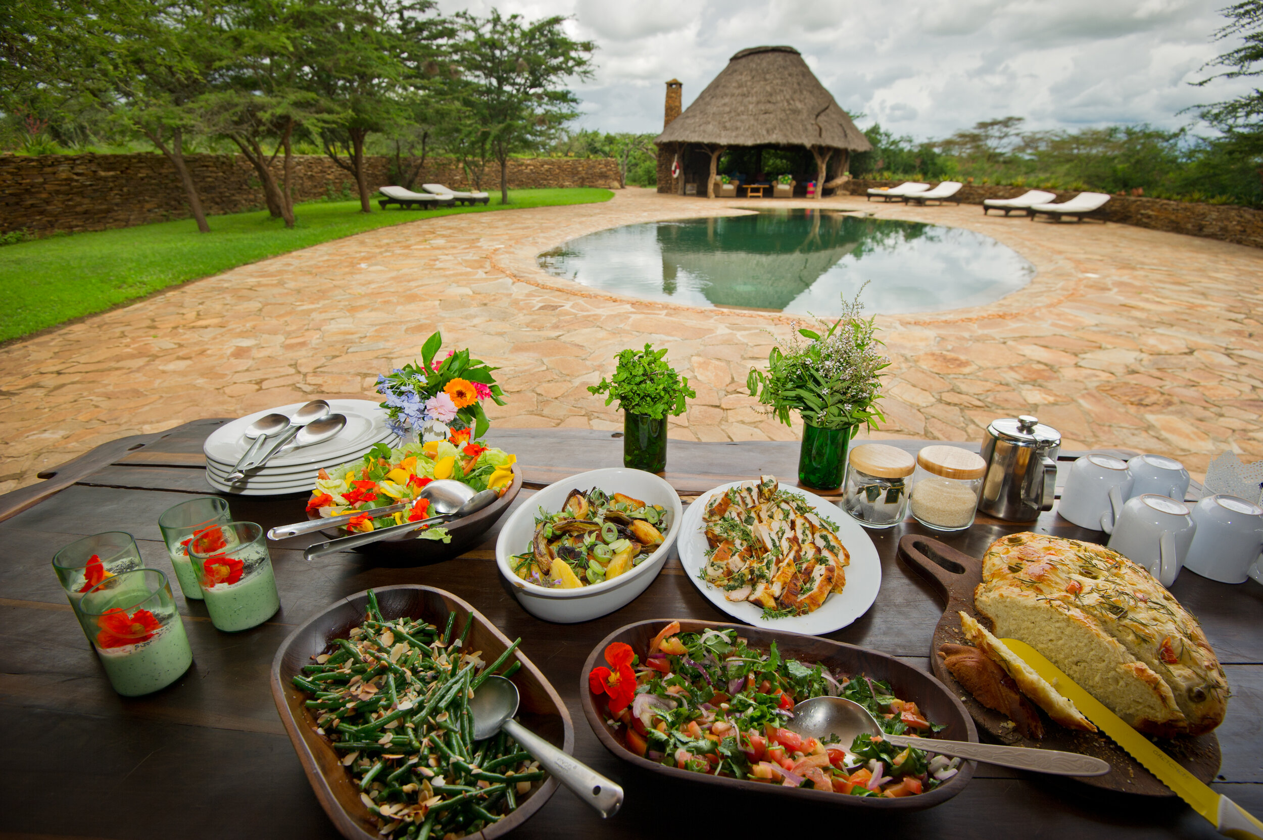 Outdoor dining table with various salads, bread, and drinks, set against a stone patio with a small pool, thatched hut, lounge chairs, and trees in the background.