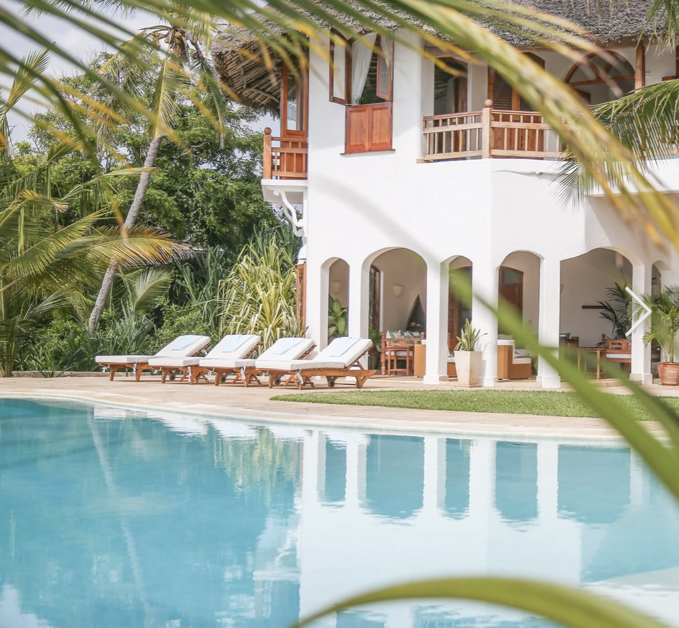 A tropical resort with a white building, outdoor pool, and sun loungers, surrounded by lush green palm trees.