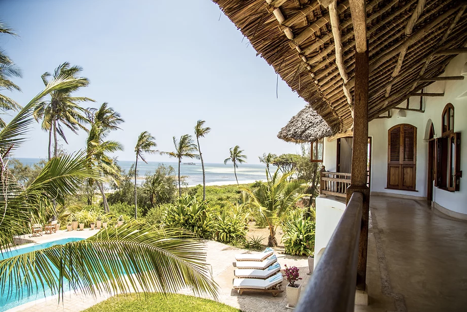Balcony view overlooking a tropical beach with palm trees, a swimming pool, and lounge chairs, featuring thatched roofs and lush greenery.