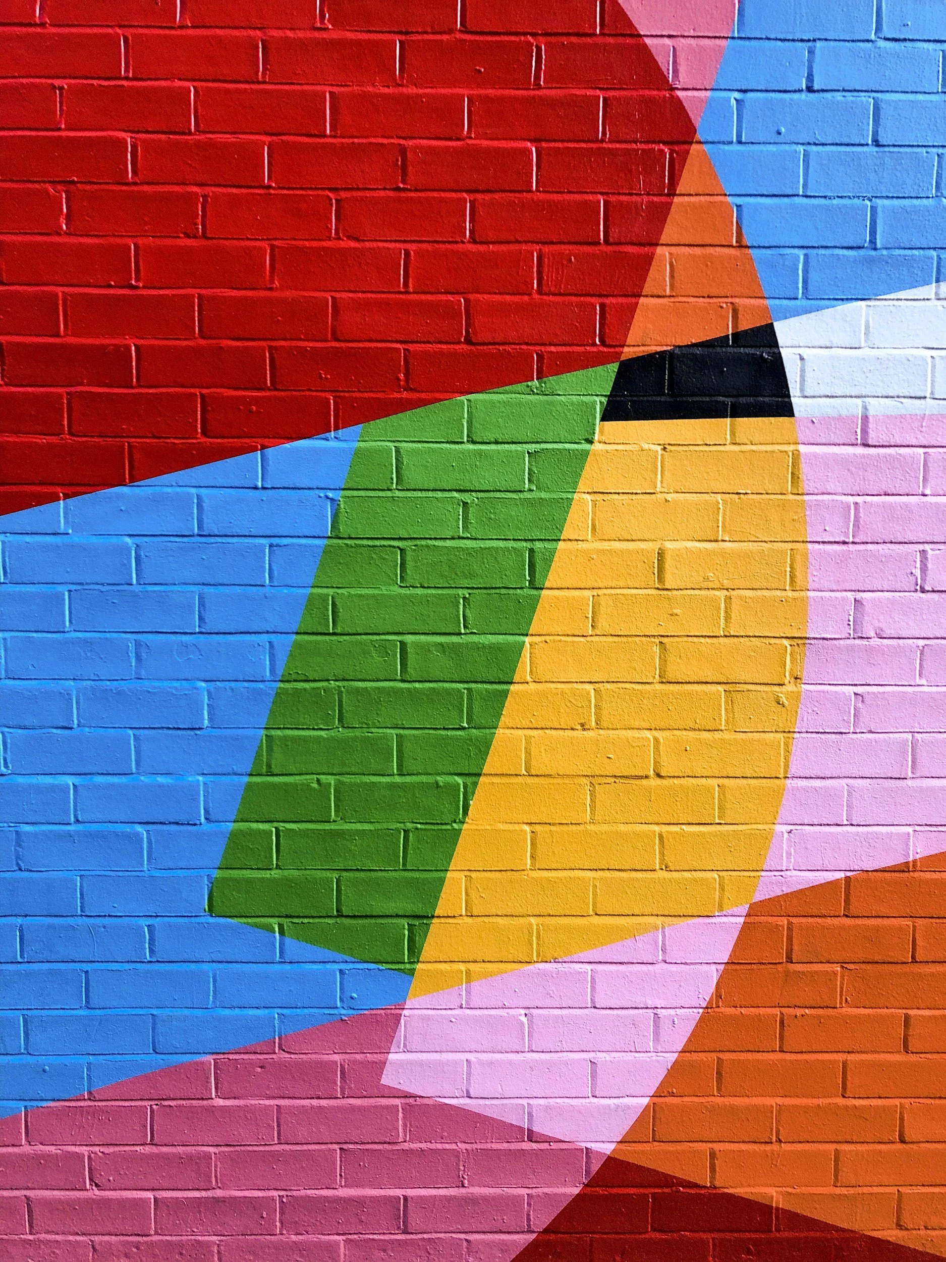 Colorful abstract geometric mural on a brick wall with overlapping shapes in red, blue, green, yellow, pink, black, and white.