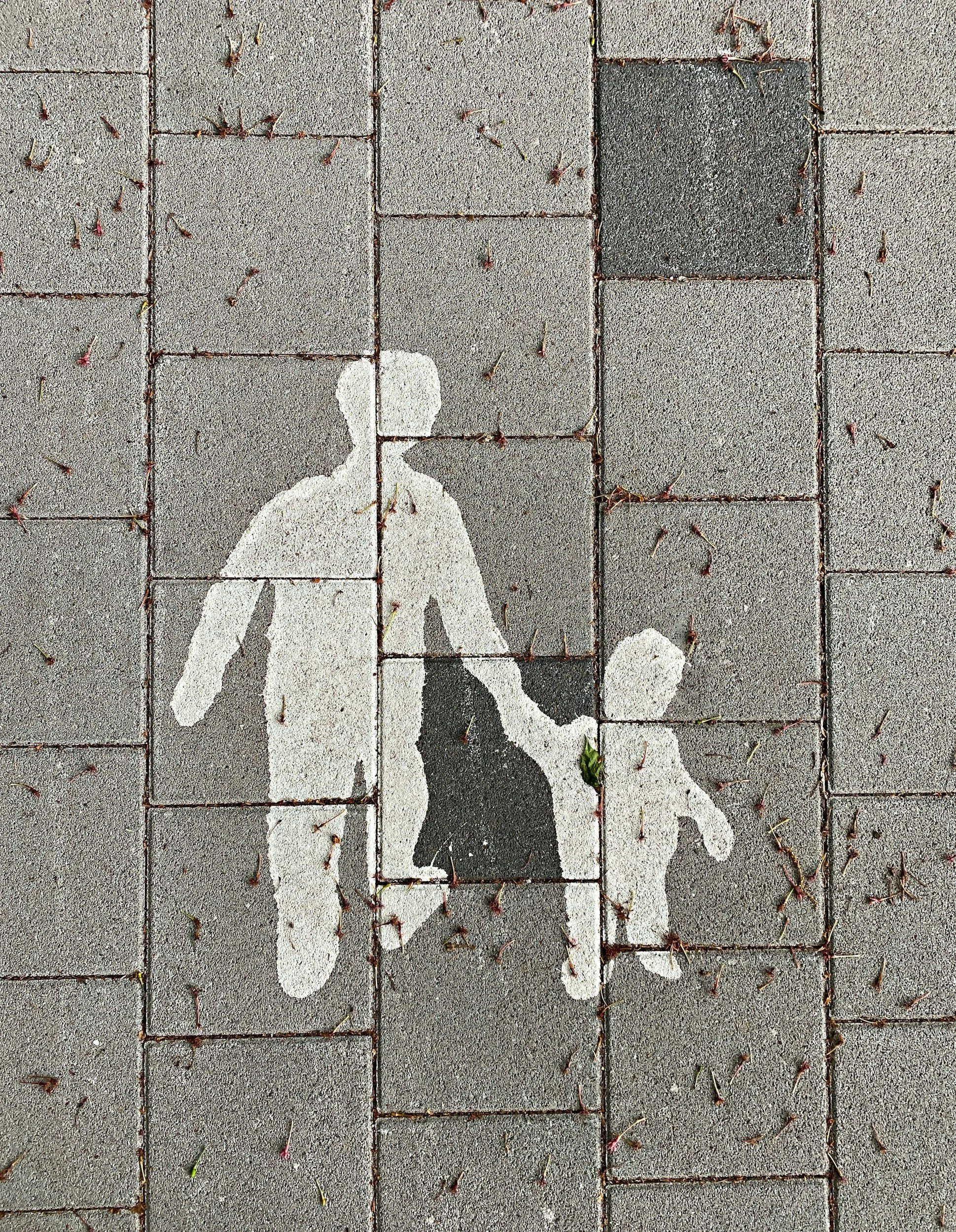 A sidewalk with a painted white silhouette of an adult holding a child's hand, both facing to the right.