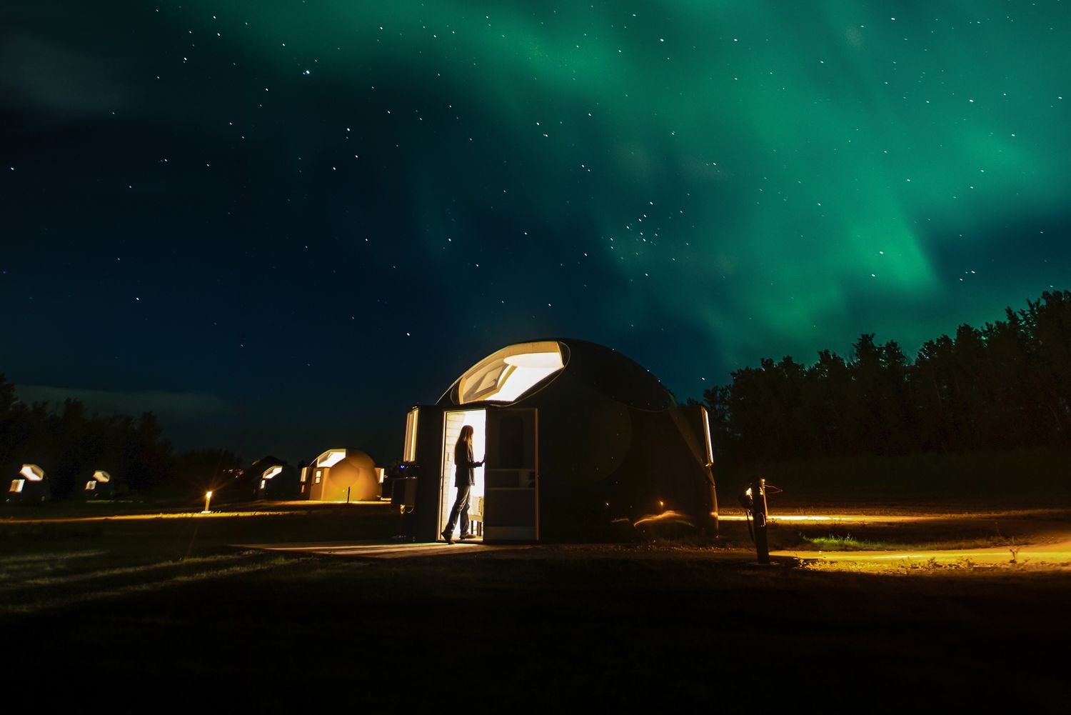 A person standing outside a glowing dome-shaped pod at night with the Northern Lights in the sky.