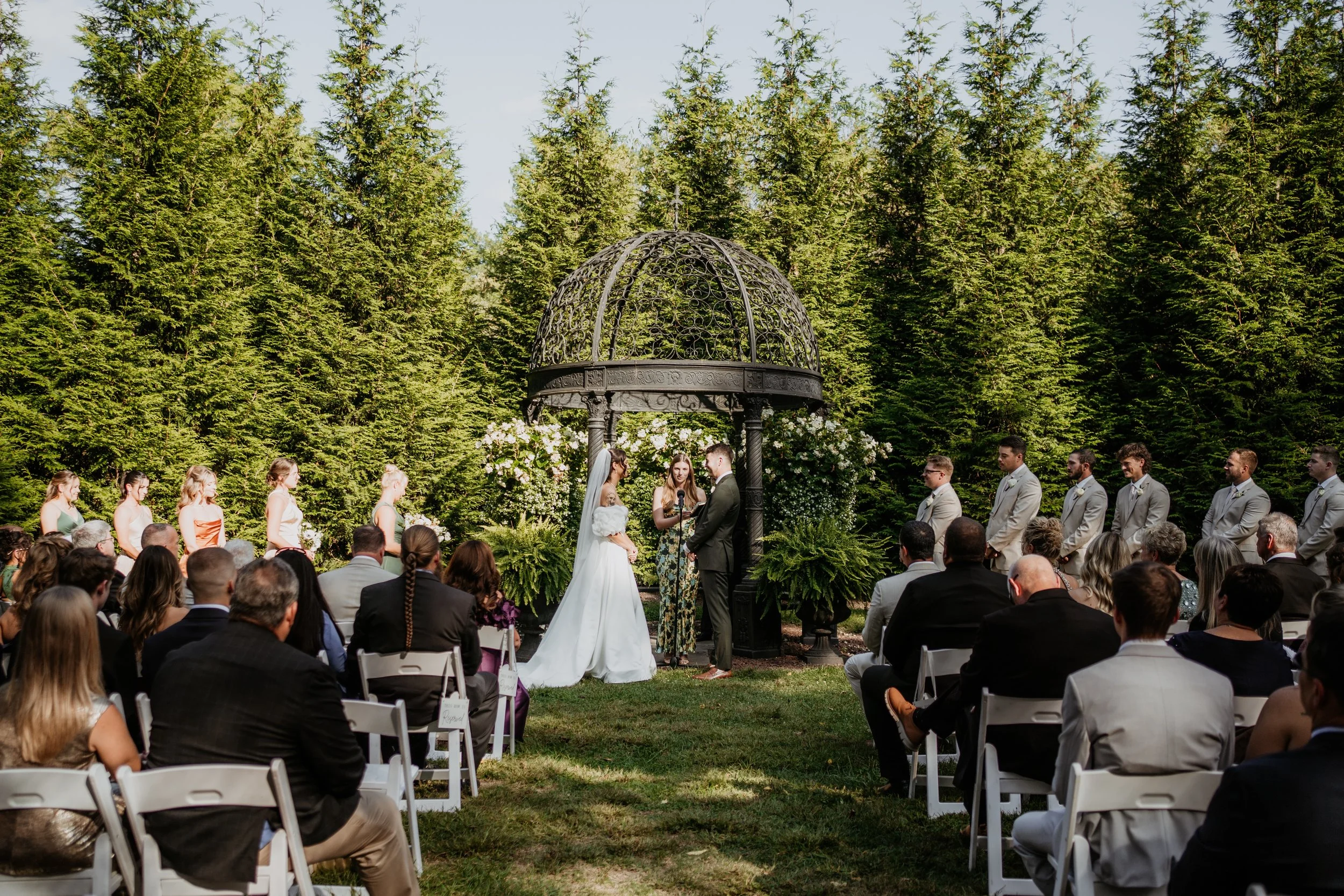a wedding ceremony with a iron gazebo and trees in the background and guests watching in chairs