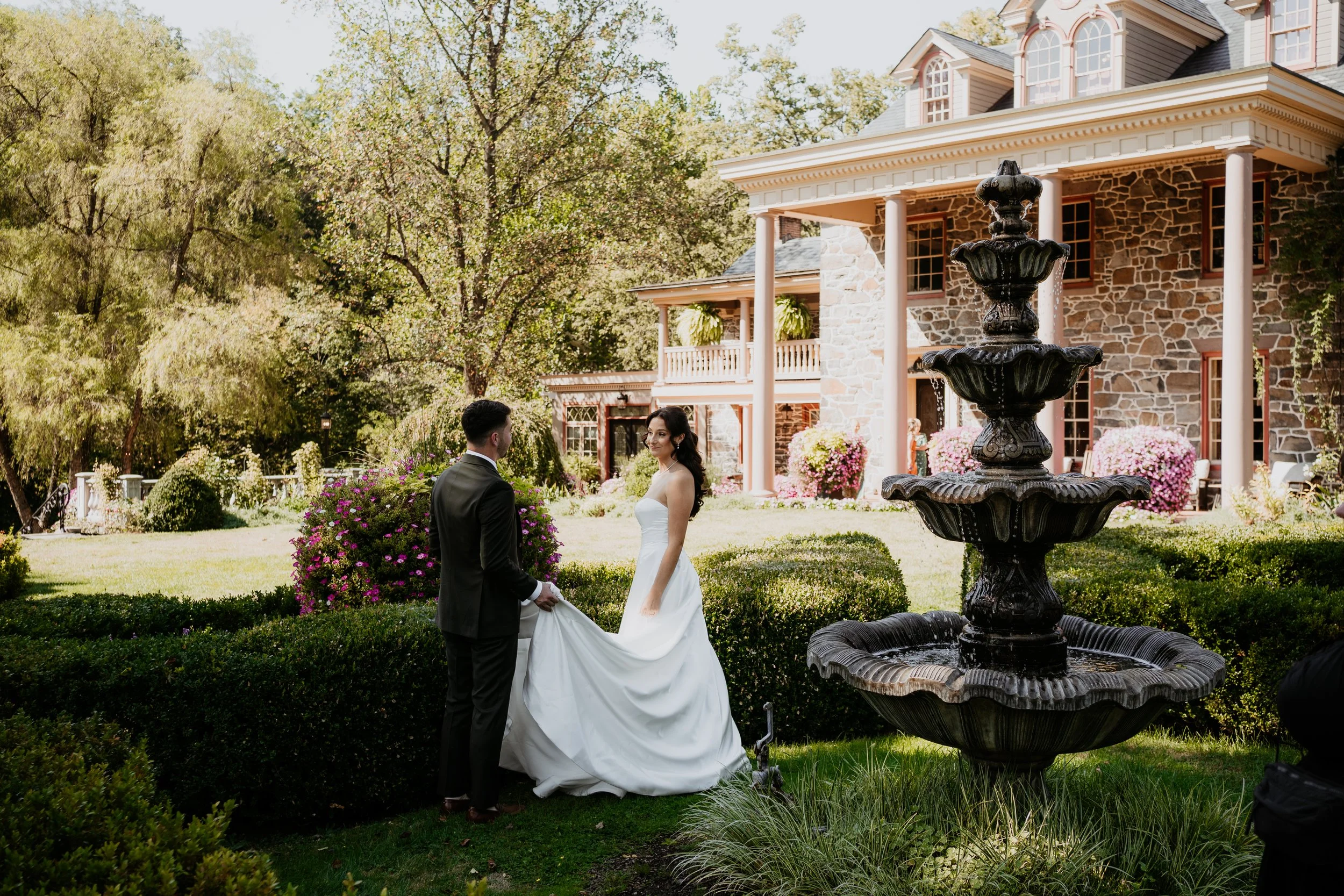 groom holds train of brides dress as they gaze at each other in the gardens of moonstone manor