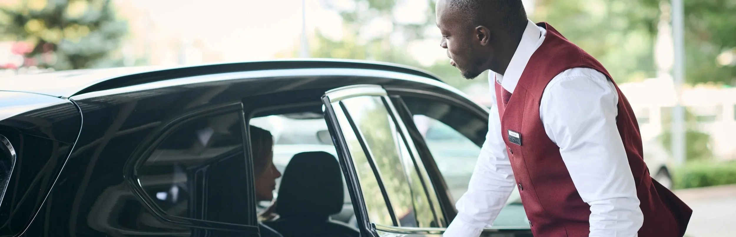 A man wearing a red vest and white shirt leaning into the front passenger side of a black car, with trees and buildings blurred in the background.