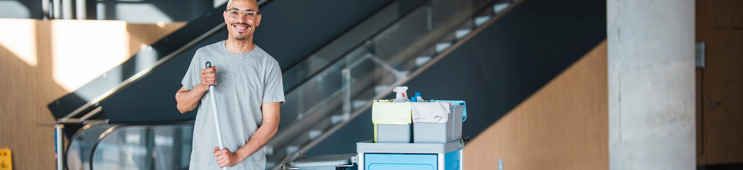 A smiling man in a gray t-shirt holding a mop next to a cleaning cart in a modern building.