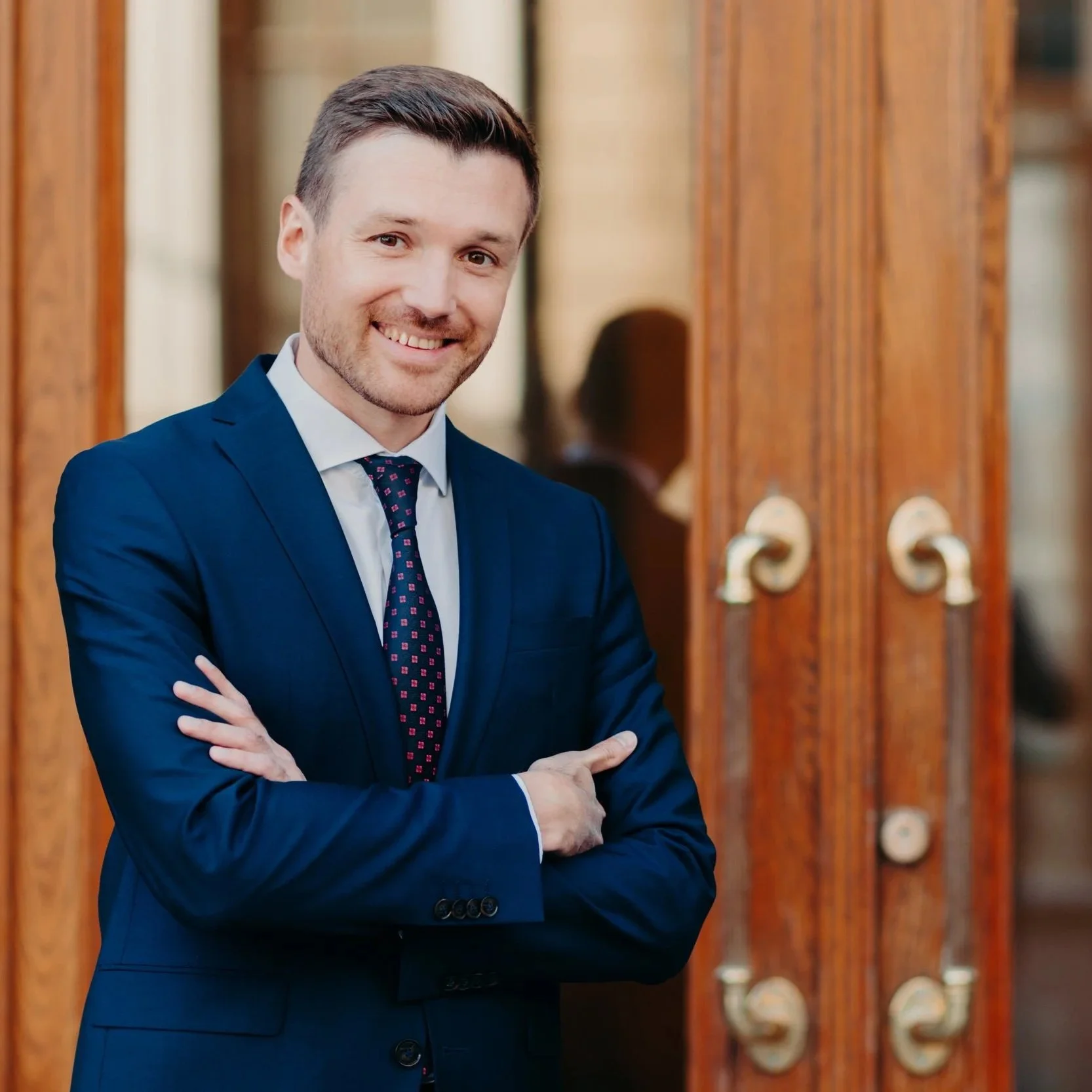 A man in a navy blue suit and tie standing in front of wooden doors, smiling with arms crossed.