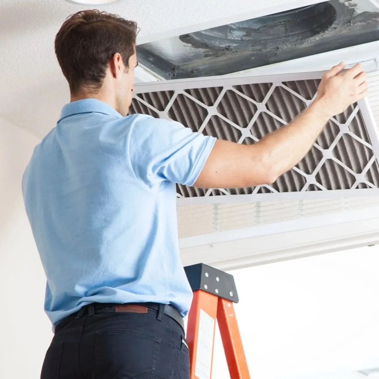 A man in a blue shirt and black pants is on a ladder, cleaning or inspecting a ceiling air conditioning unit filter.