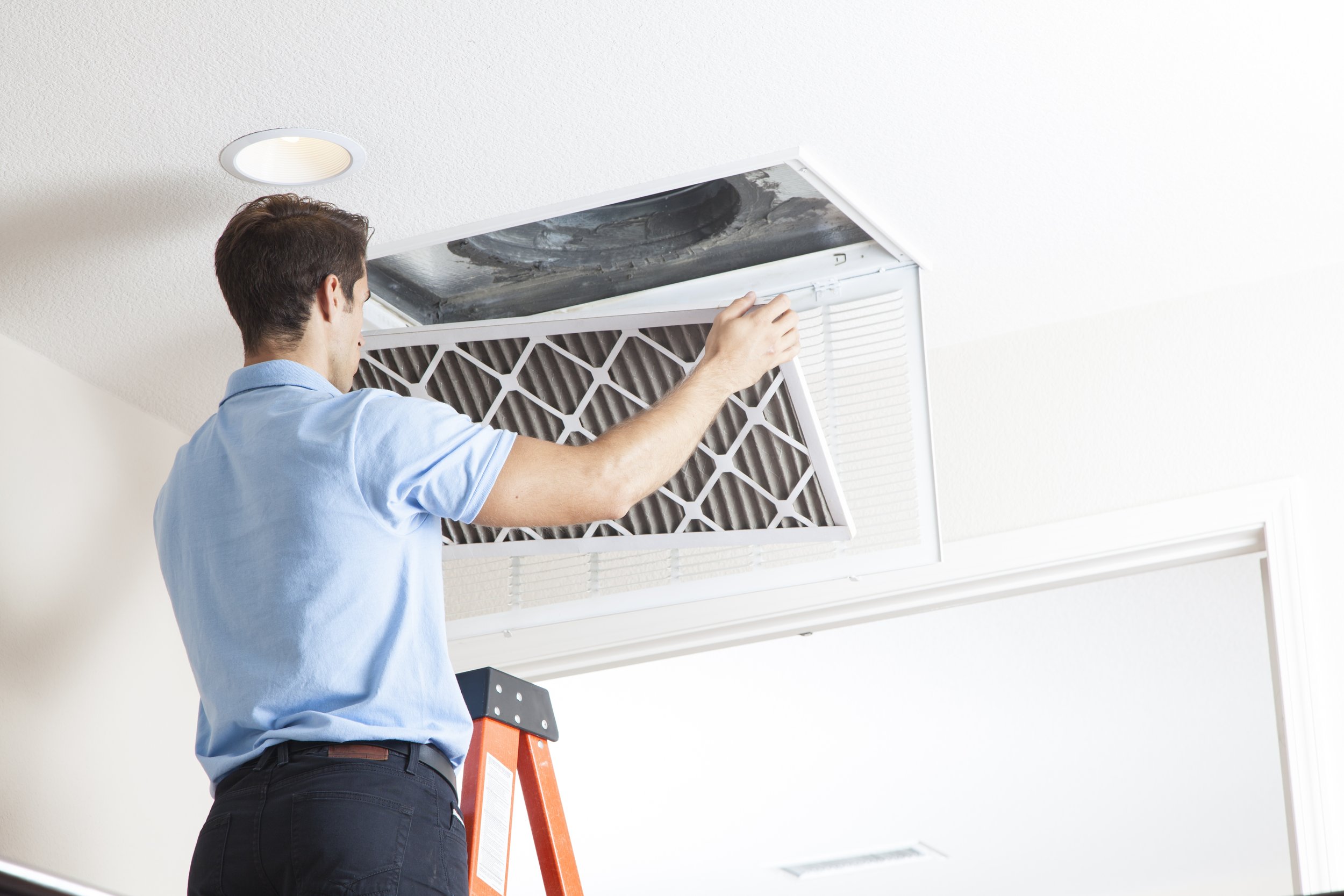 A man in a light blue shirt standing on an orange ladder, removing a dirty air filter from a ceiling-mounted air conditioning unit.
