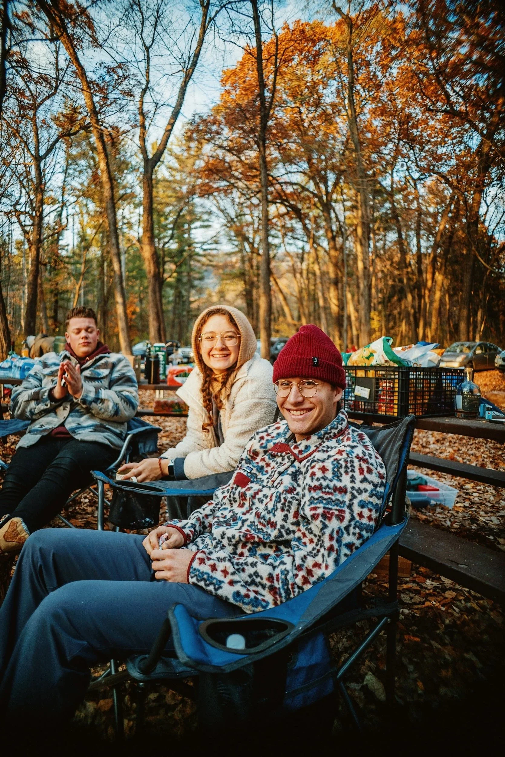 Three smiling young adults sitting outdoors in a wooded park during autumn, with fallen leaves on the ground and trees with orange and yellow leaves in the background, enjoying a picnic.