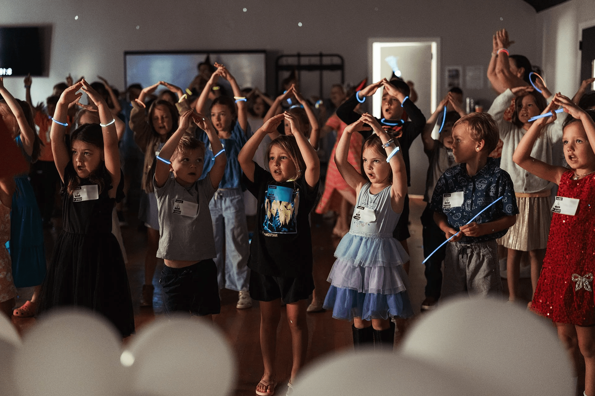 Children dancing and playing with glow sticks at a celebration or party, viewed through a pattern of white circles.