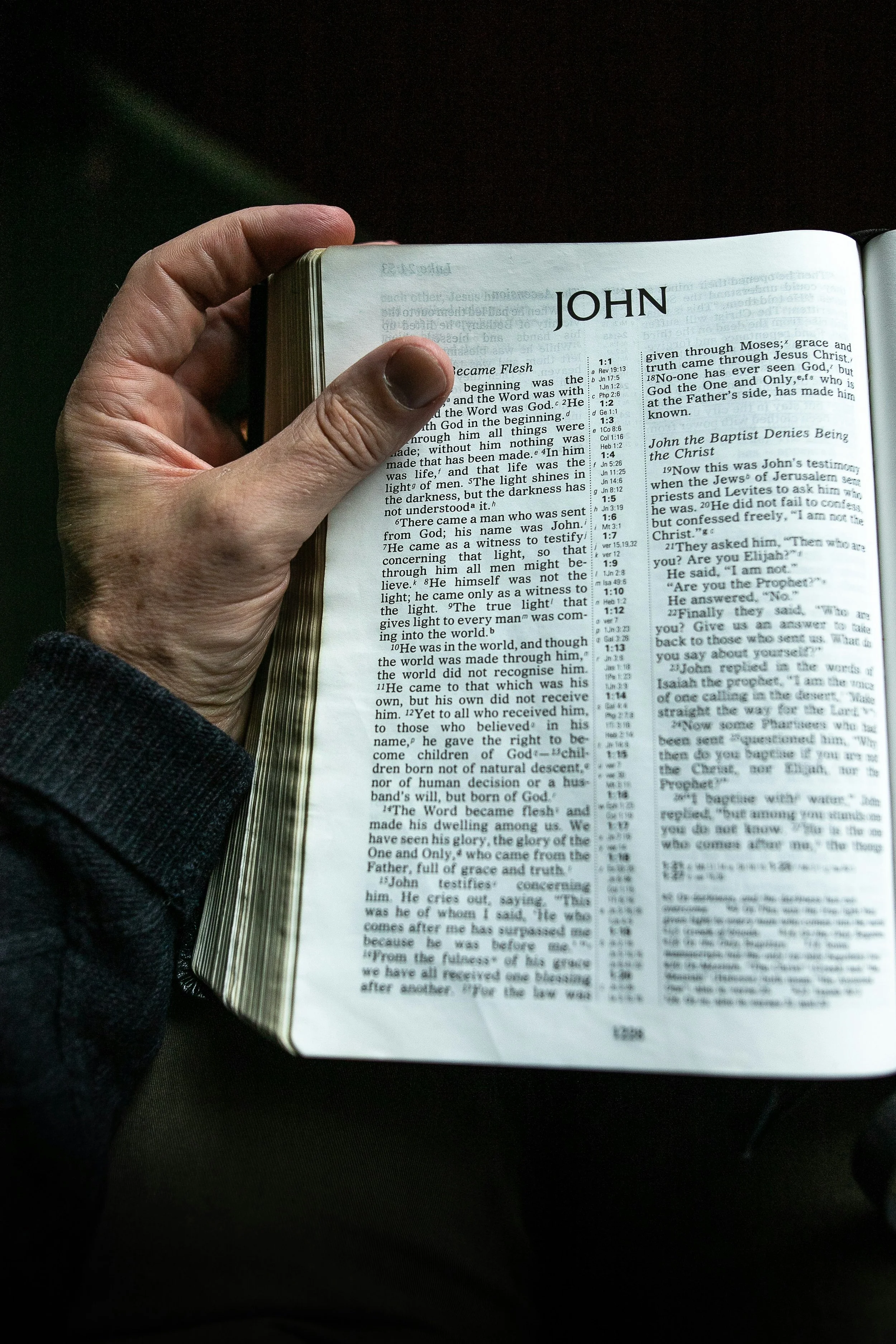 A person holding an open Bible with the page titled "JOHN" in large font. The Bible page contains text from the New Testament, including references and verses.