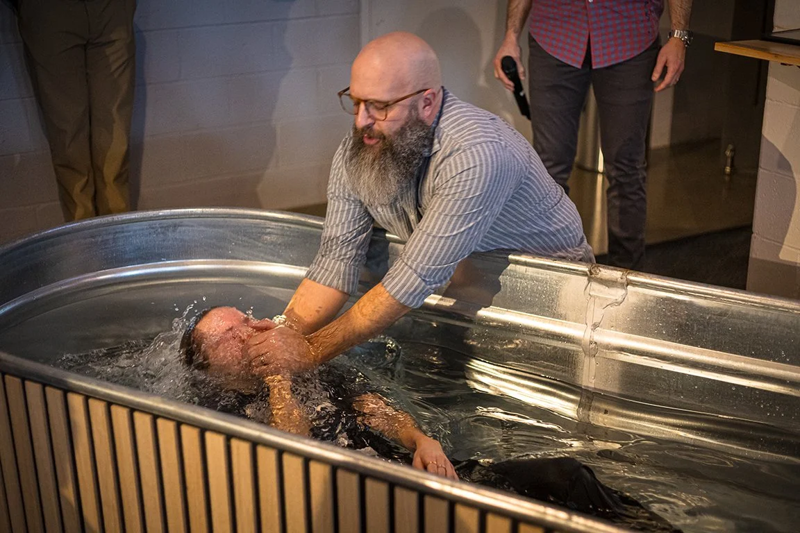 A man with a beard and glasses is baptizing another person in a large metal baptismal pool.