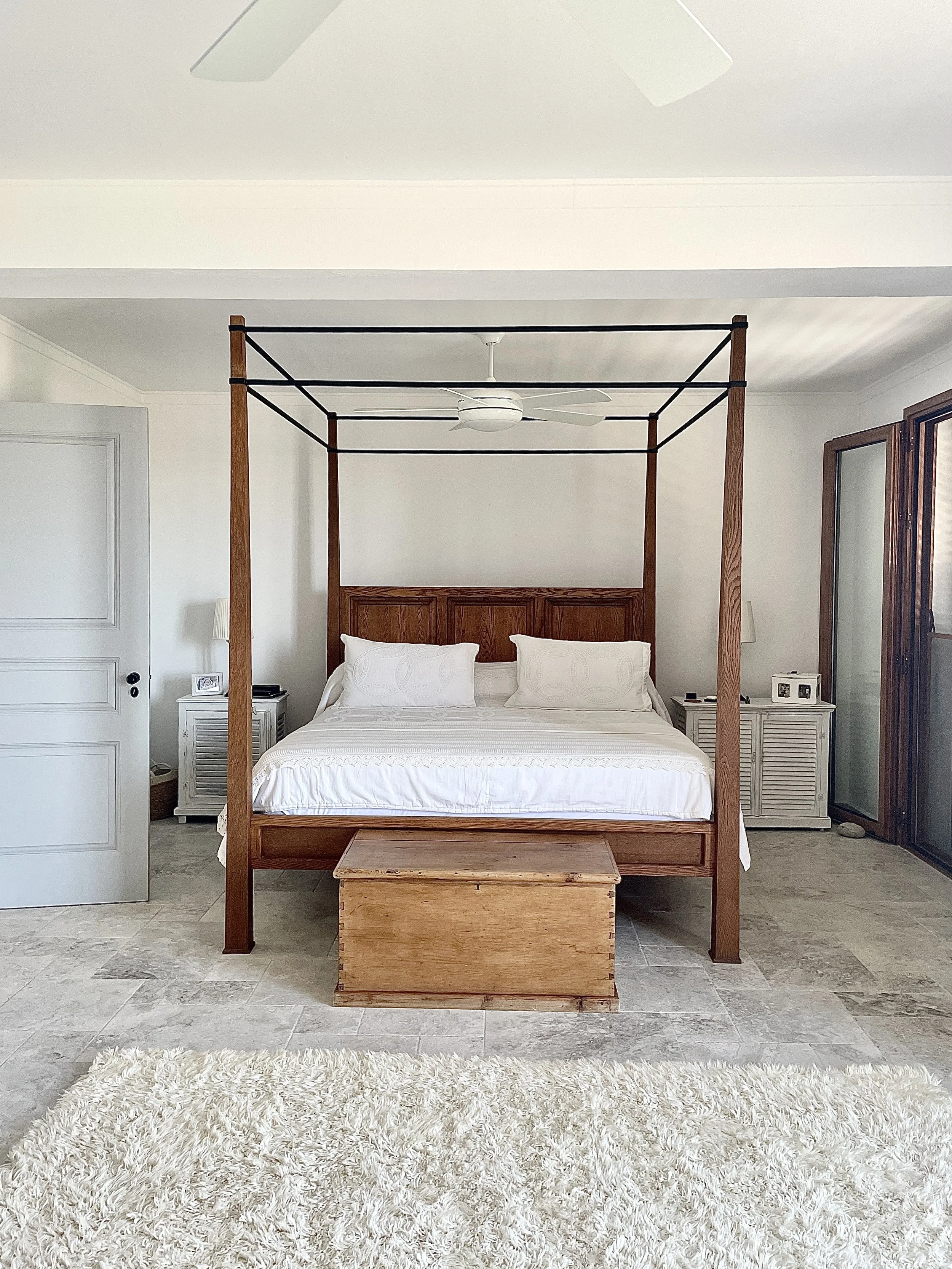 A bedroom with a four-poster bed, white bedding, and bedside tables on either side, with a ceiling fan overhead and wooden framed sliding doors to the right.
