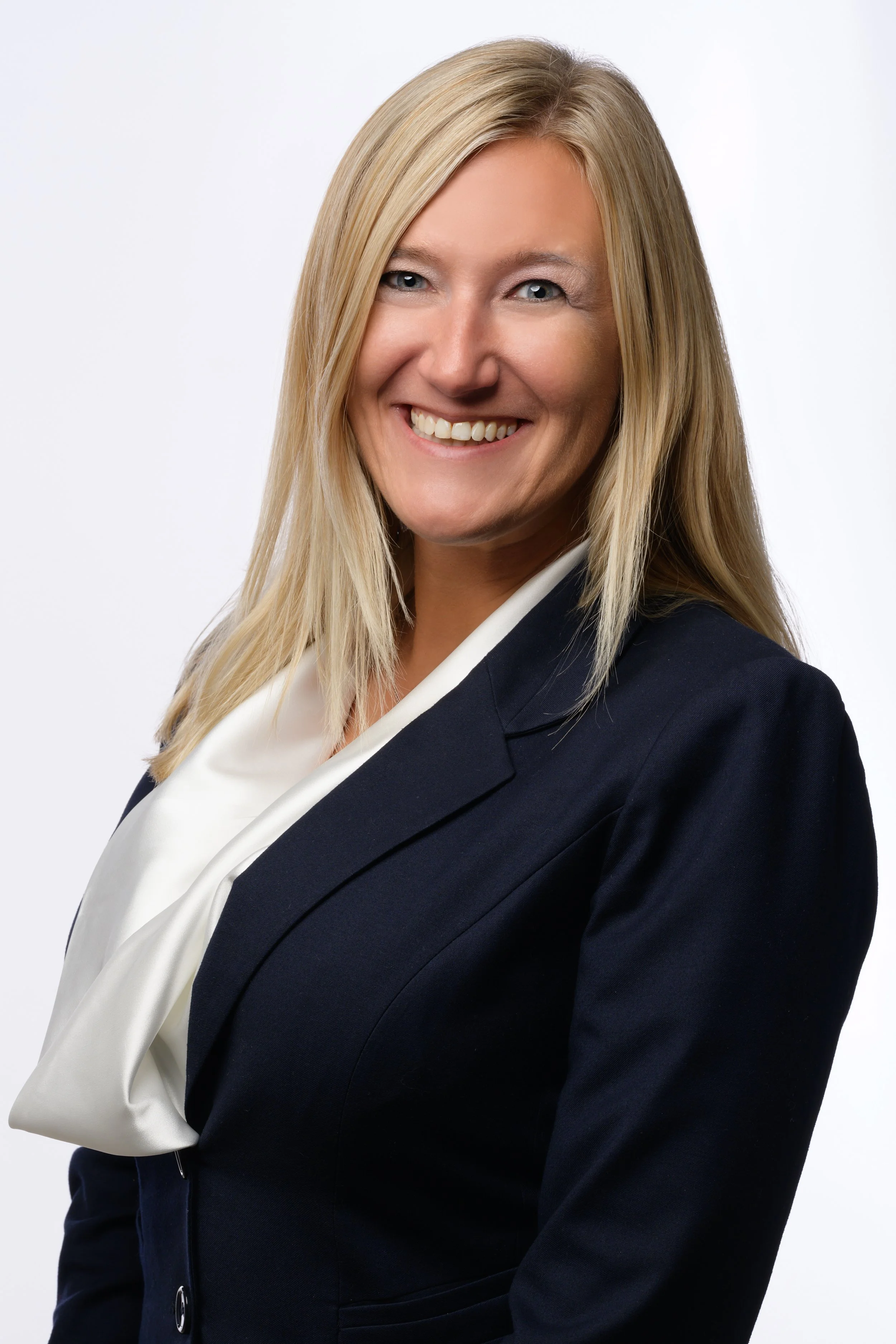 A smiling woman with blonde hair wearing a black blazer and white blouse, posing against a plain white background.