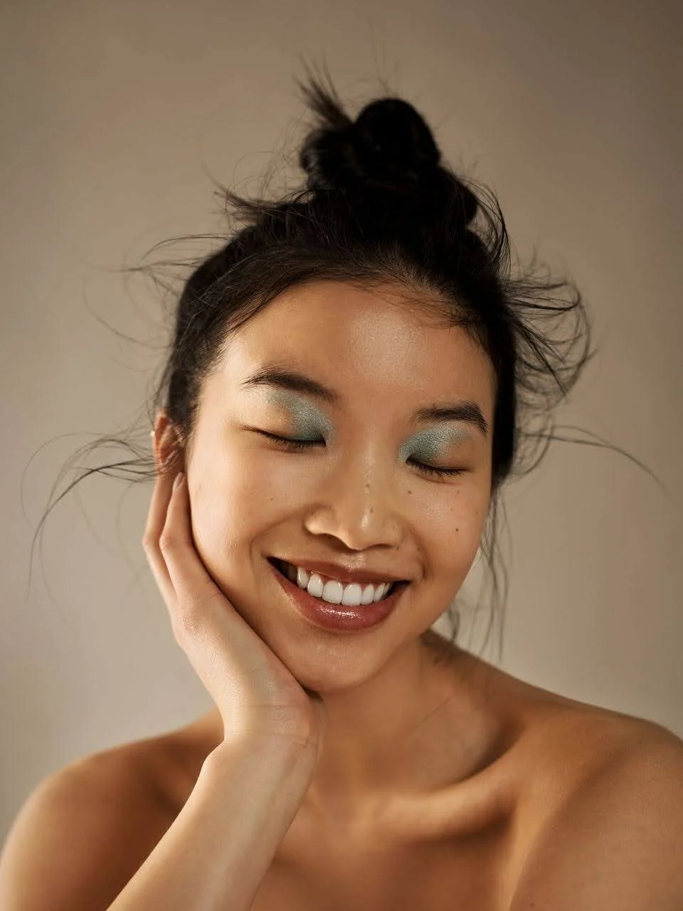 Close-up of a young woman with black hair styled in a bun, smiling with closed eyes, touching her face gently, wearing light blue eyeshadow and no shirt.