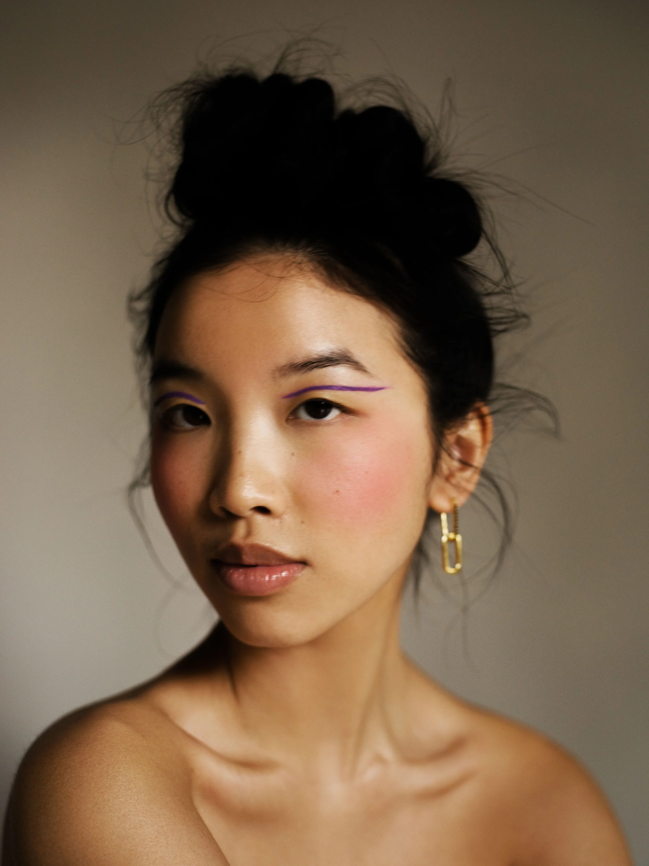 Portrait of a woman with dark hair styled in a messy bun, wearing gold earrings, with makeup including purple eyeliner, pink blush, and natural lips, against a neutral background.