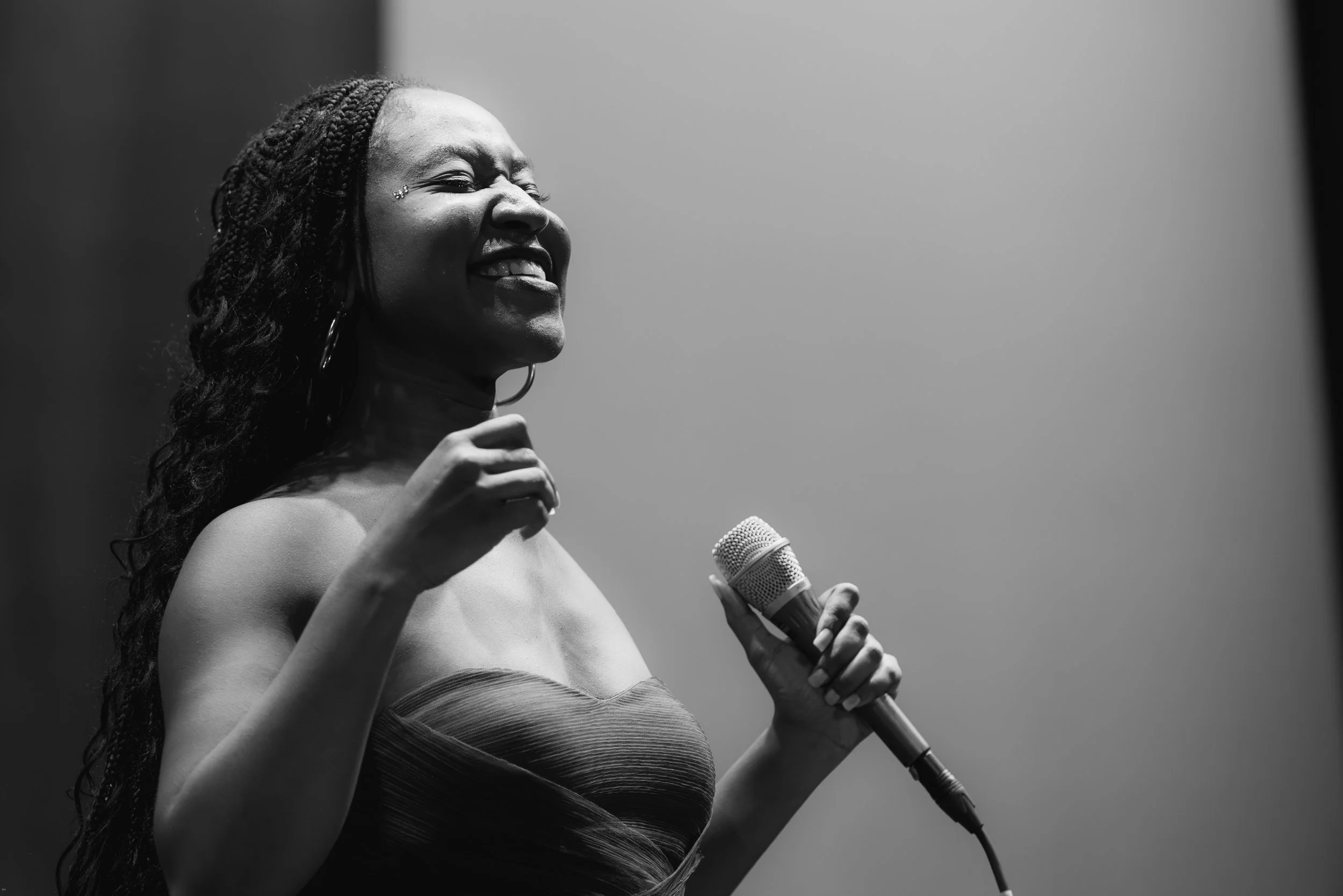 A black and white photo of a woman speaking into a microphone, smiling, with long braided hair, wearing a strapless top.