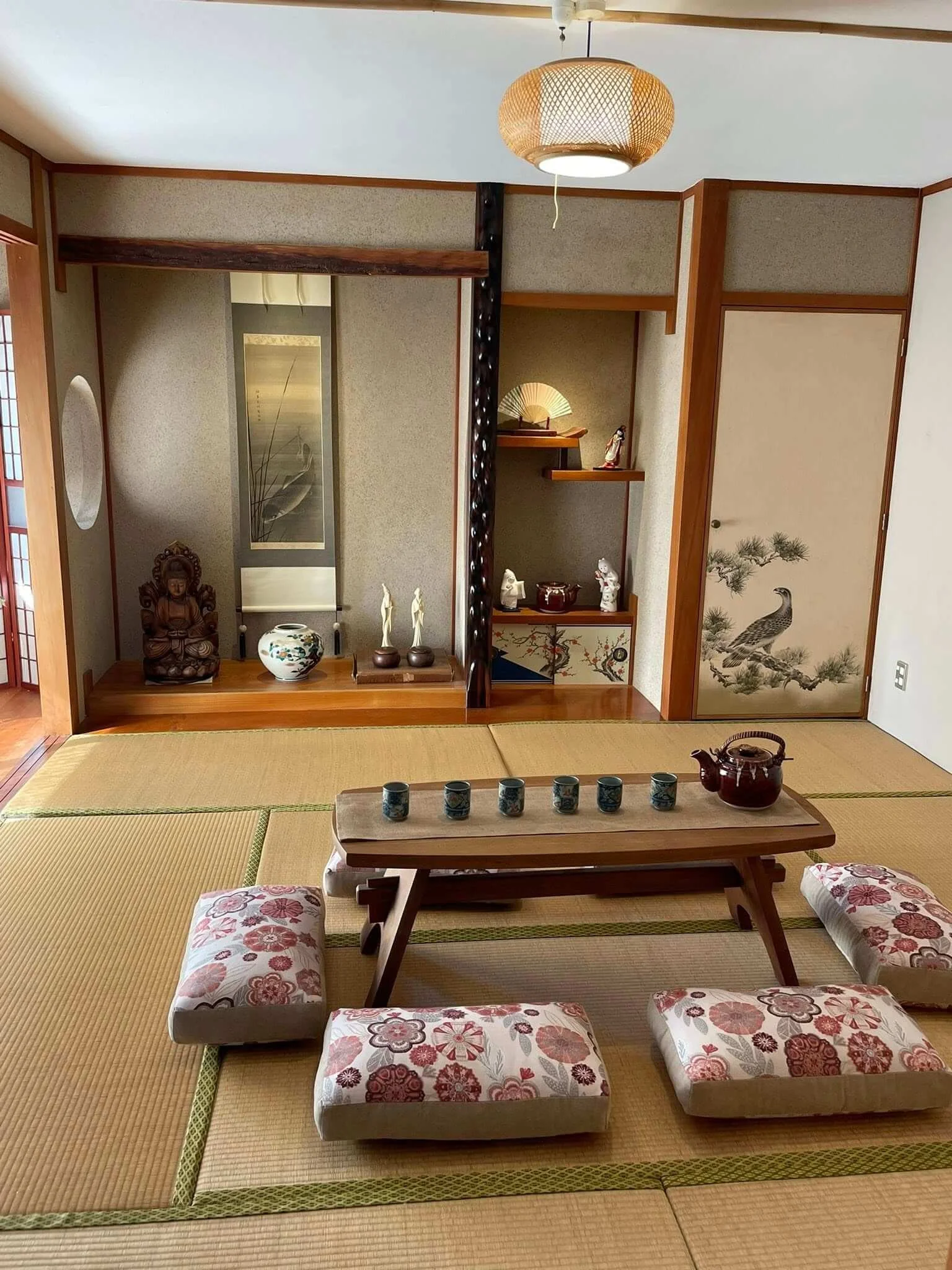 A traditional Japanese tatami room with a low wooden table, four floor cushions, and decorative items on shelves, including ceramics and a fan, with sliding doors featuring a peacock and pine tree painting.
