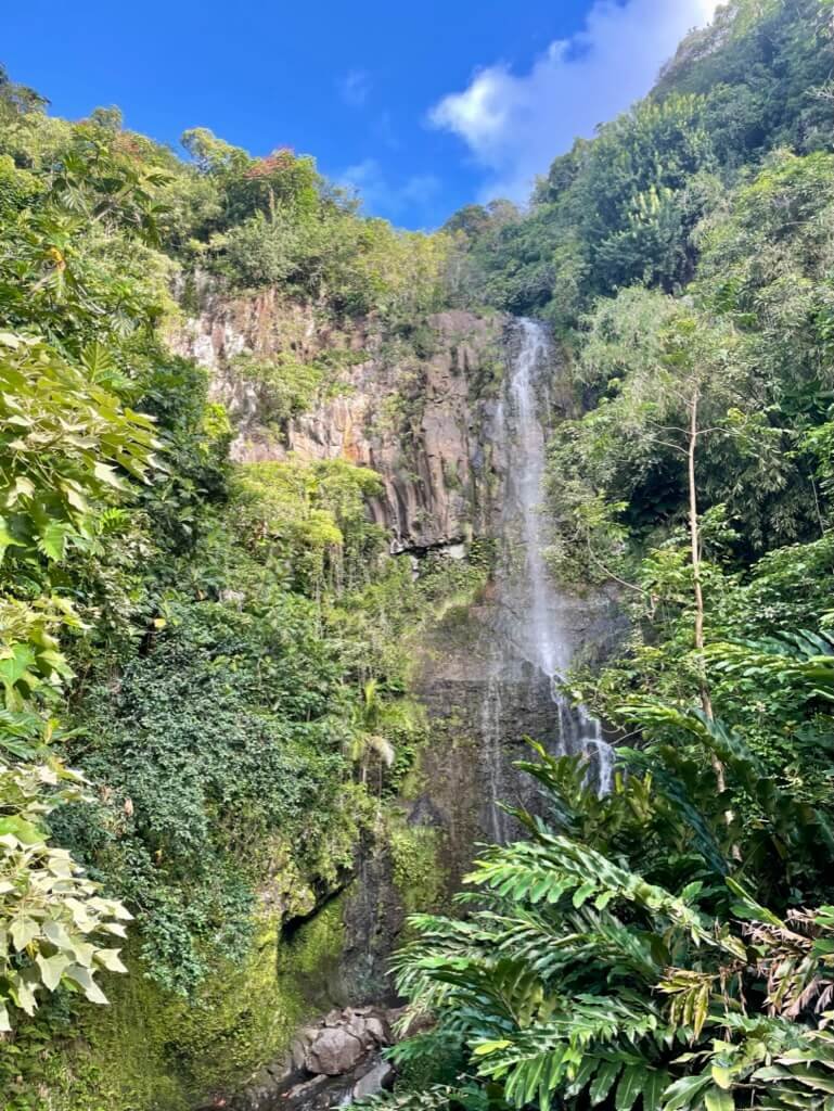 Lush green tropical forest surrounding a tall thin waterfall cascading down a cliff into rocks below, with a bright blue sky and some white clouds overhead.