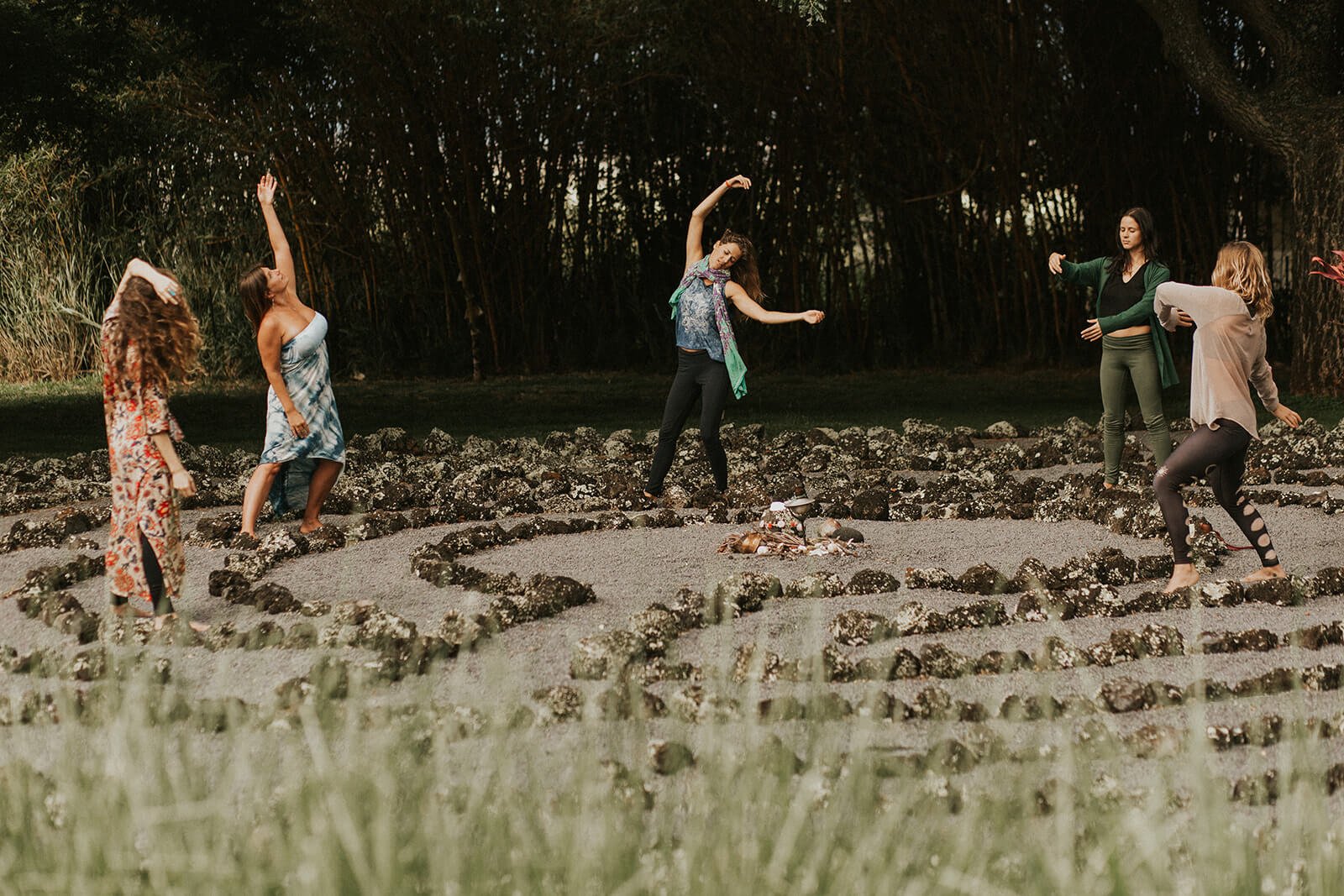 Six women dancing around a fire circle outdoors, with tall trees in the background, during daylight.