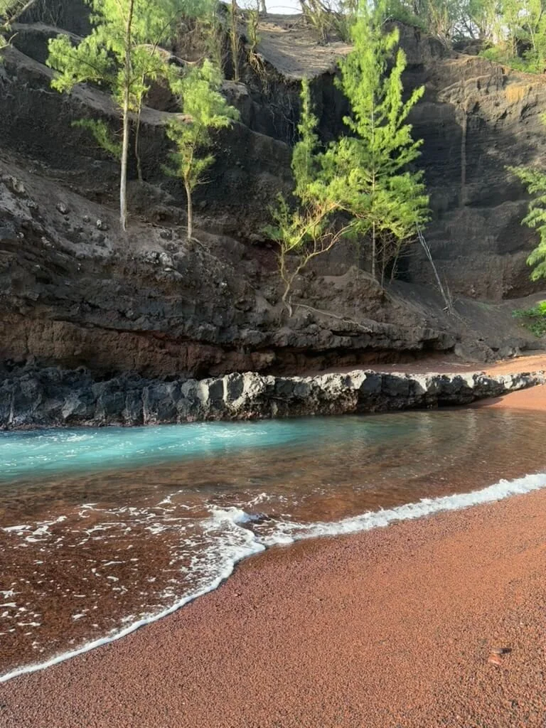 A small beach with reddish sand, a stream of clear blue water, rocky cliffs, and green trees.