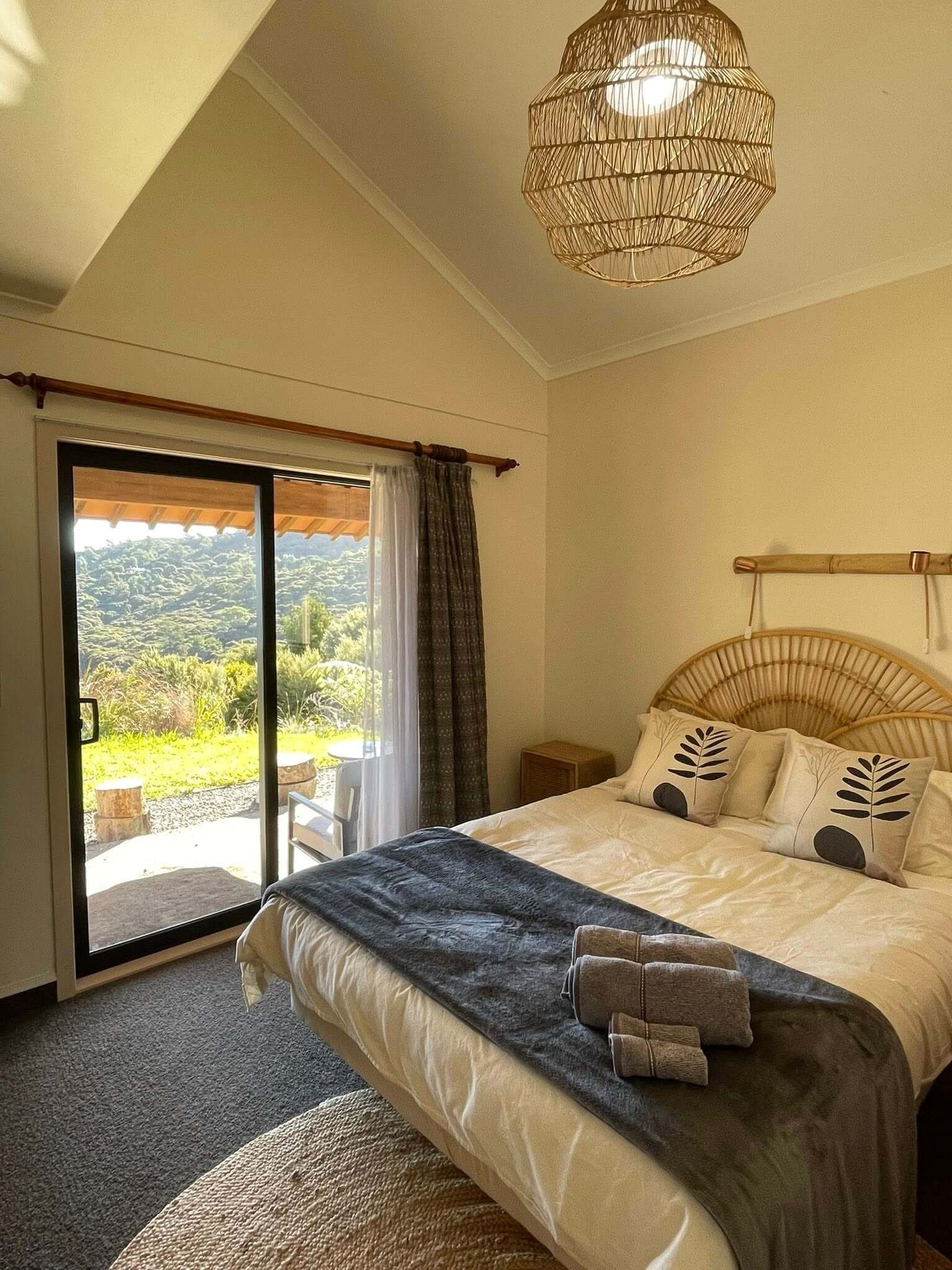 A bedroom with a bed featuring a leaf-patterned pillow, a dark gray blanket, and a beige cover. There are rolled towels on the bed. To the left, an open sliding glass door leads outside, showing a lush, green landscape with trees and a hill in the distance. The room has beige walls, a textured rug, and a wooden hanging rod with curtains. A rattan ceiling light is turned on.