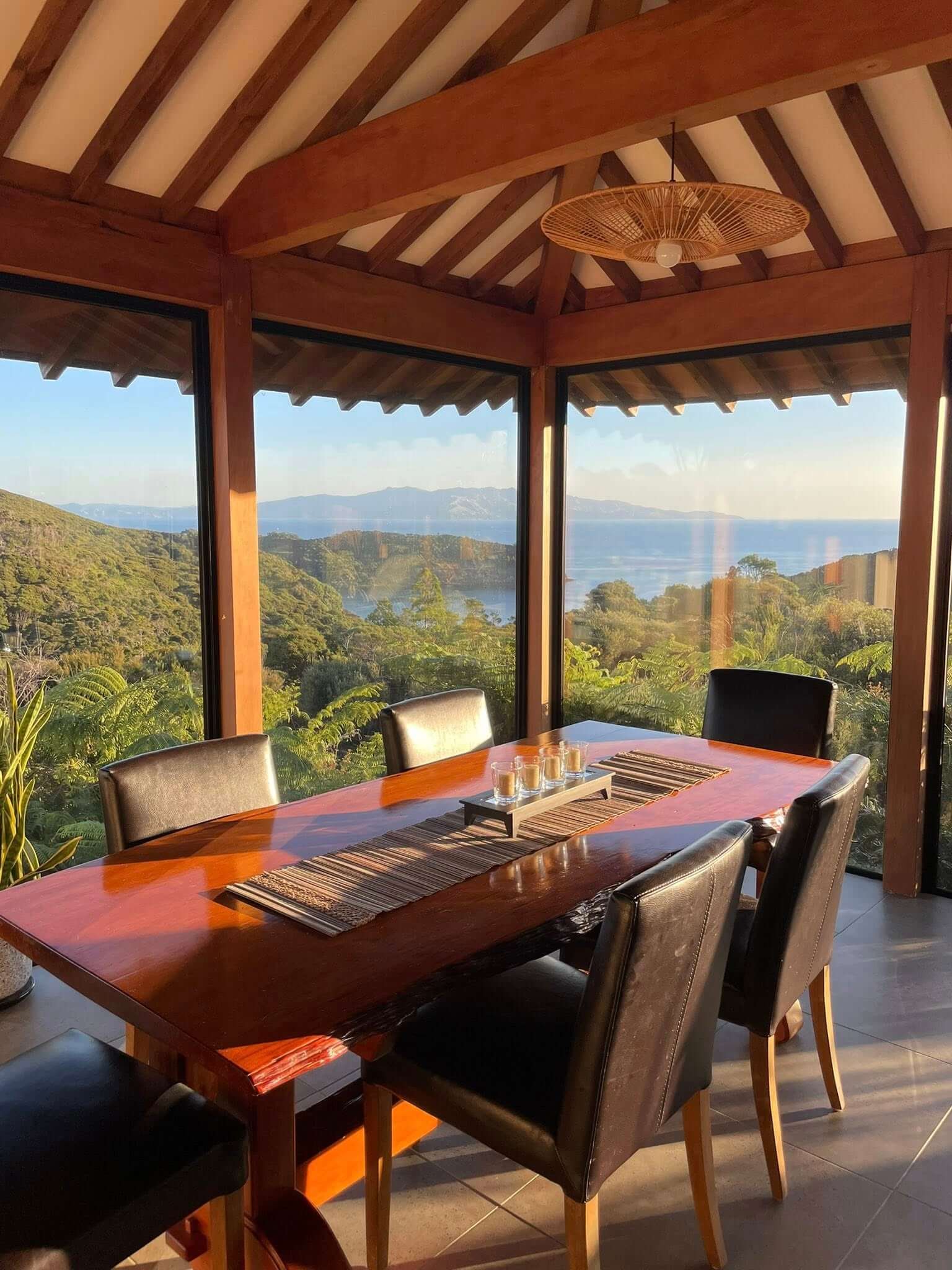 A dining room with a wooden table and six black chairs near large glass windows showing a scenic view of green hills, ocean, and distant mountains.