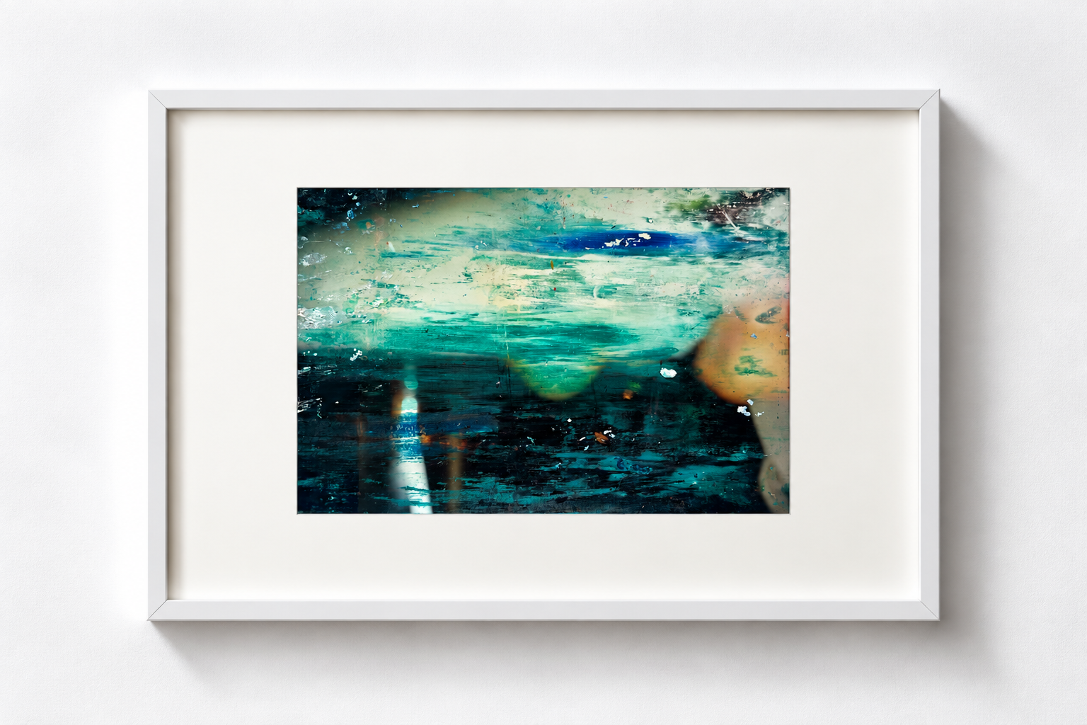 Abstract photo with shades of blue, green, beige, and black, framed in white, on a white wall.