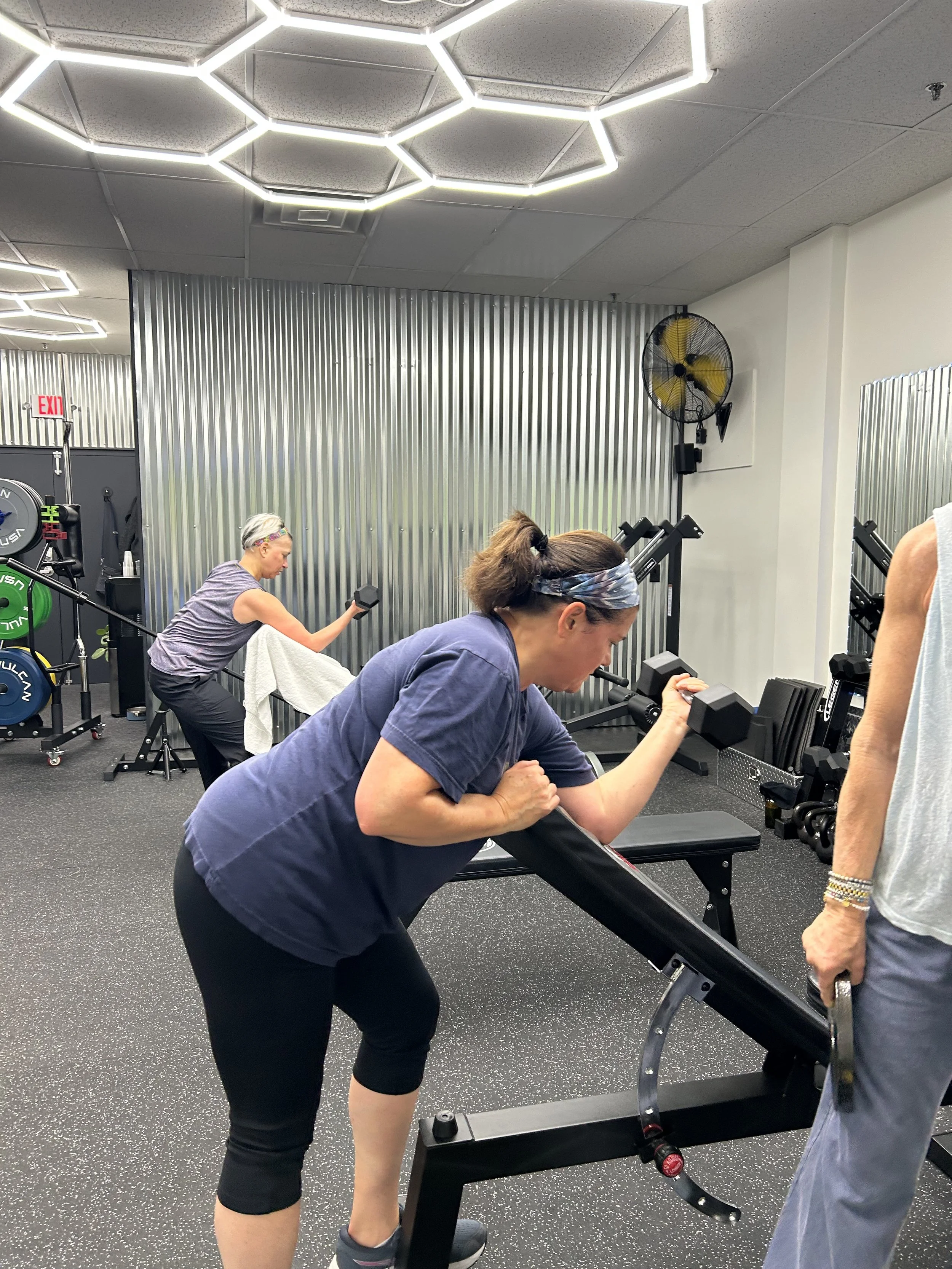 Two women and one person with a sleeveless shirt and bracelets on their arm working out in a gym. One woman is using a rowing machine, another woman is using dumbbells, and a third person is standing nearby.