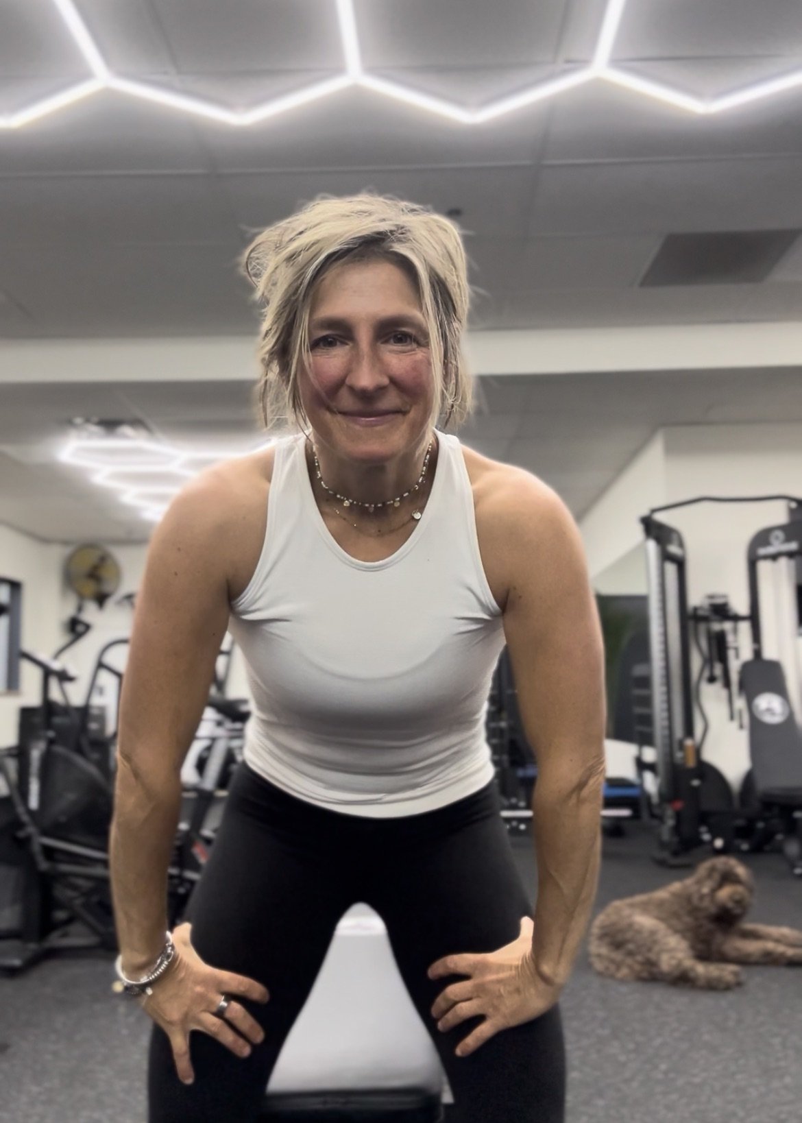 A woman with short blonde hair smiling at the camera in a gym, wearing a white tank top and black workout pants with a dog lying on the floor behind her.