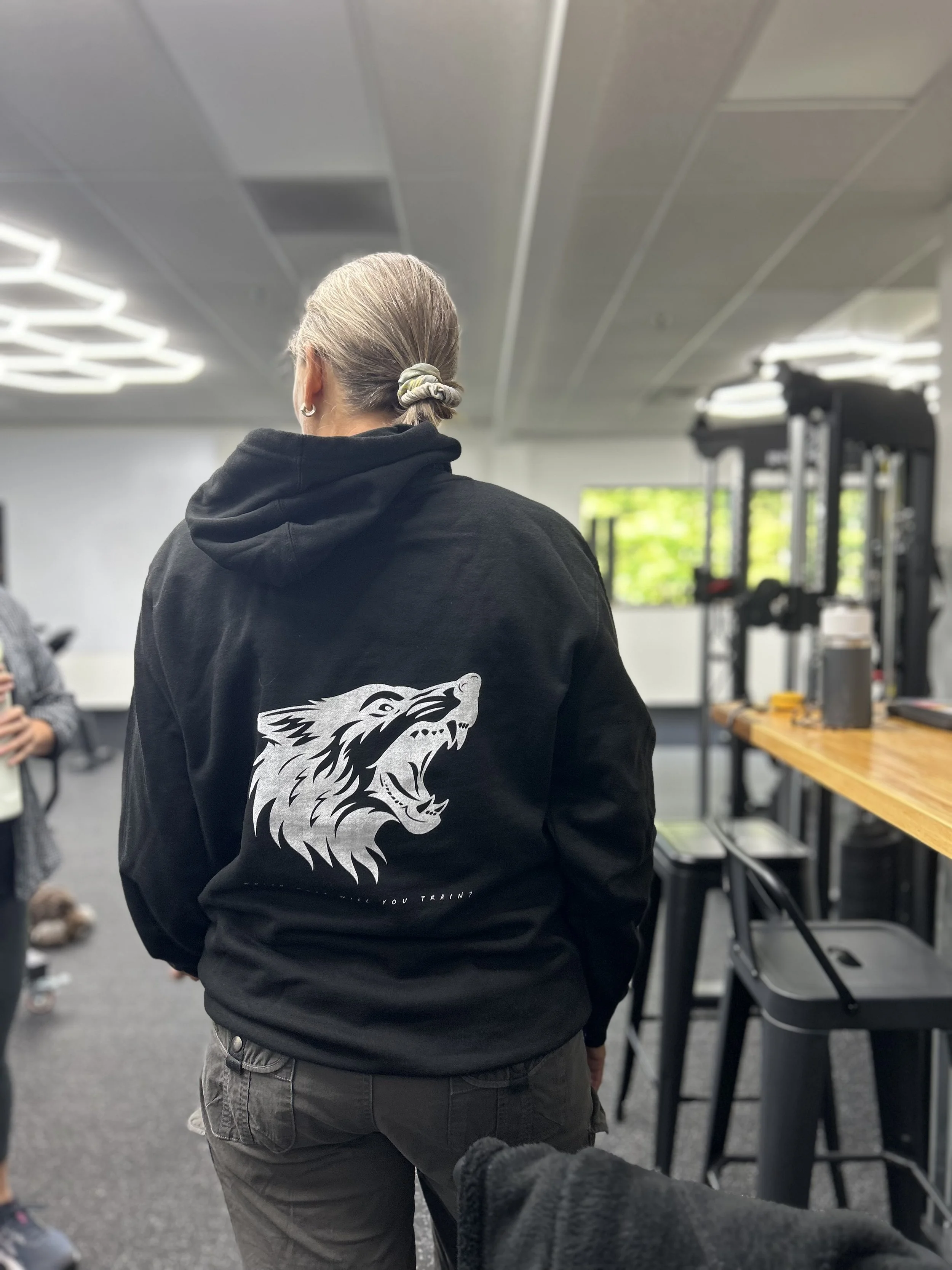 A person wearing a black hoodie with a wolf graphic on the back standing in a gym or fitness center.