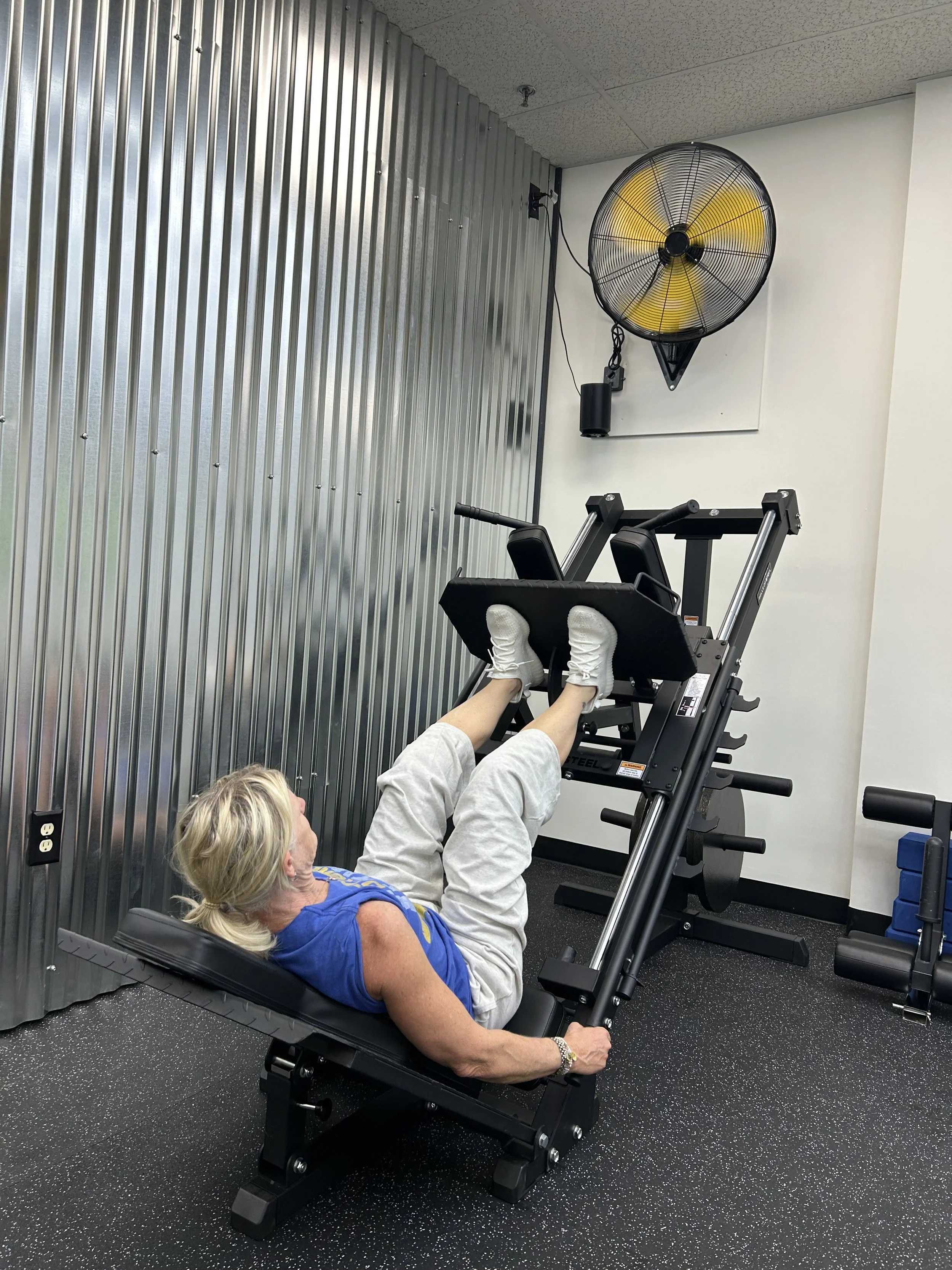 Woman exercising on a leg press machine in a gym, with a metal wall and a wall-mounted fan in the background.
