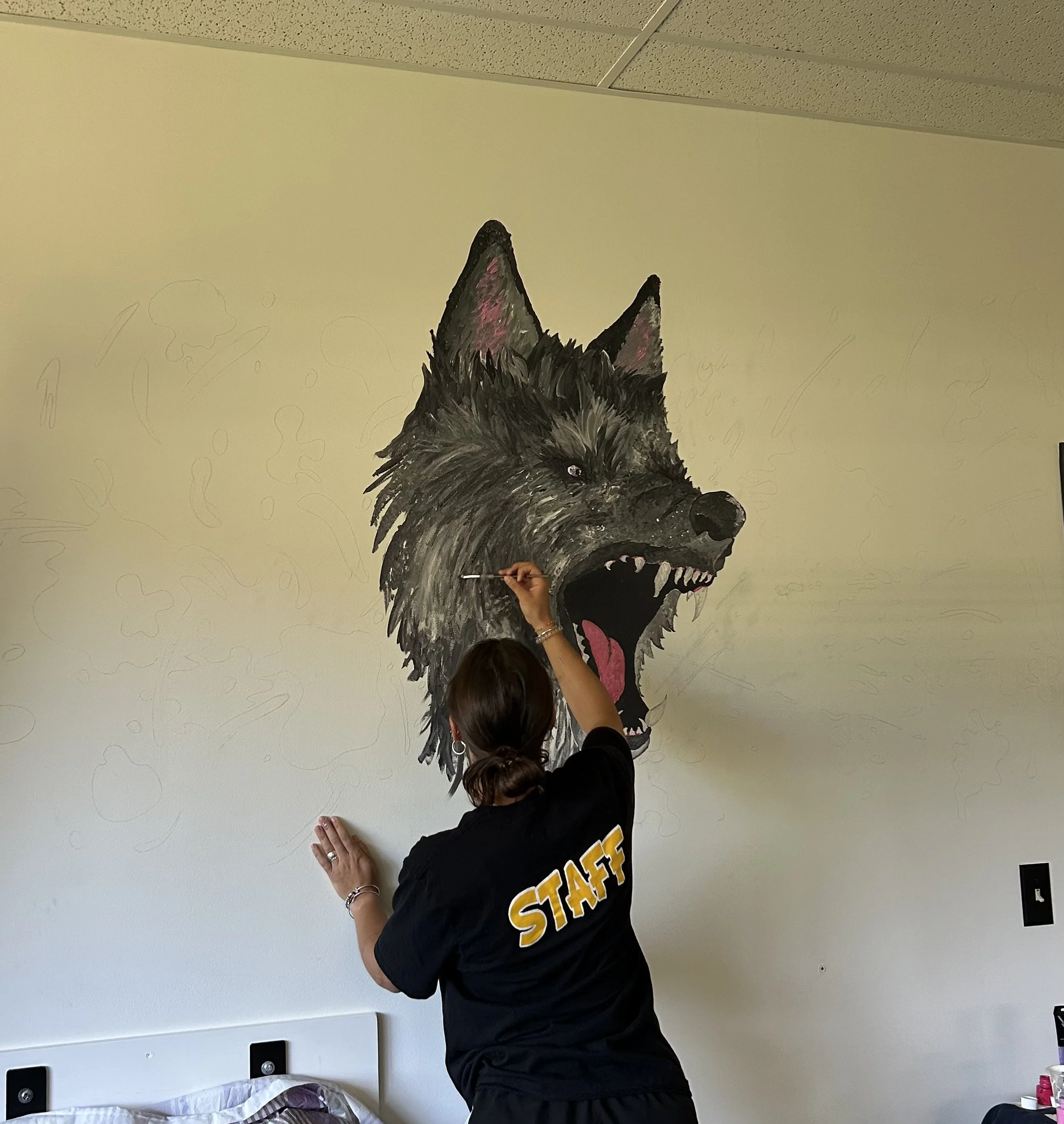 A person wearing a black shirt with the word 'STAFF' painting a large, detailed mural of a snarling wolf on a wall.