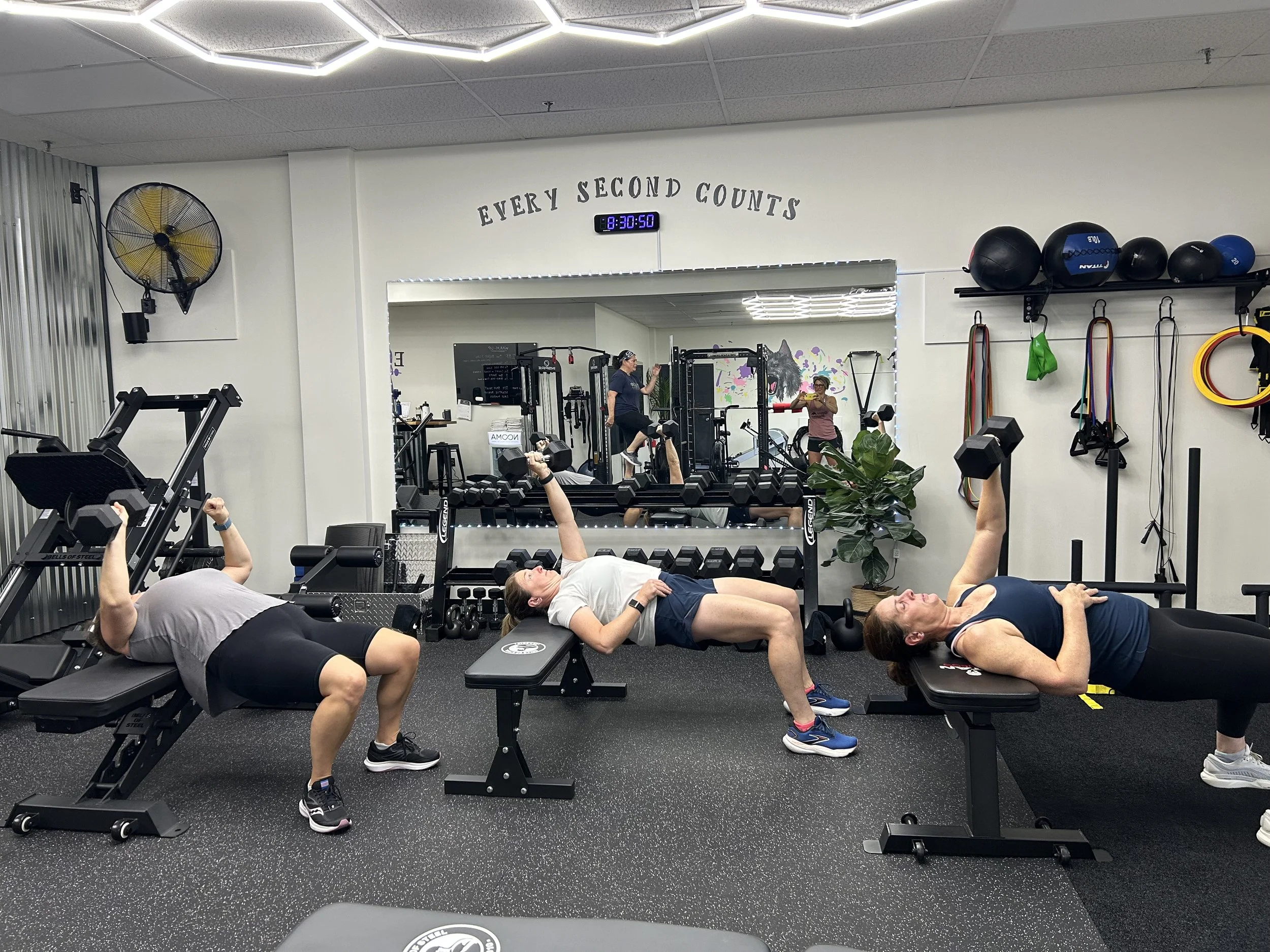 Three women exercising with dumbbells in a gym, doing exercises on benches and on the floor. Mirror reflecting the gym environment, and fitness equipment on the wall, including medicine balls, resistance bands, and gymnastic rings.