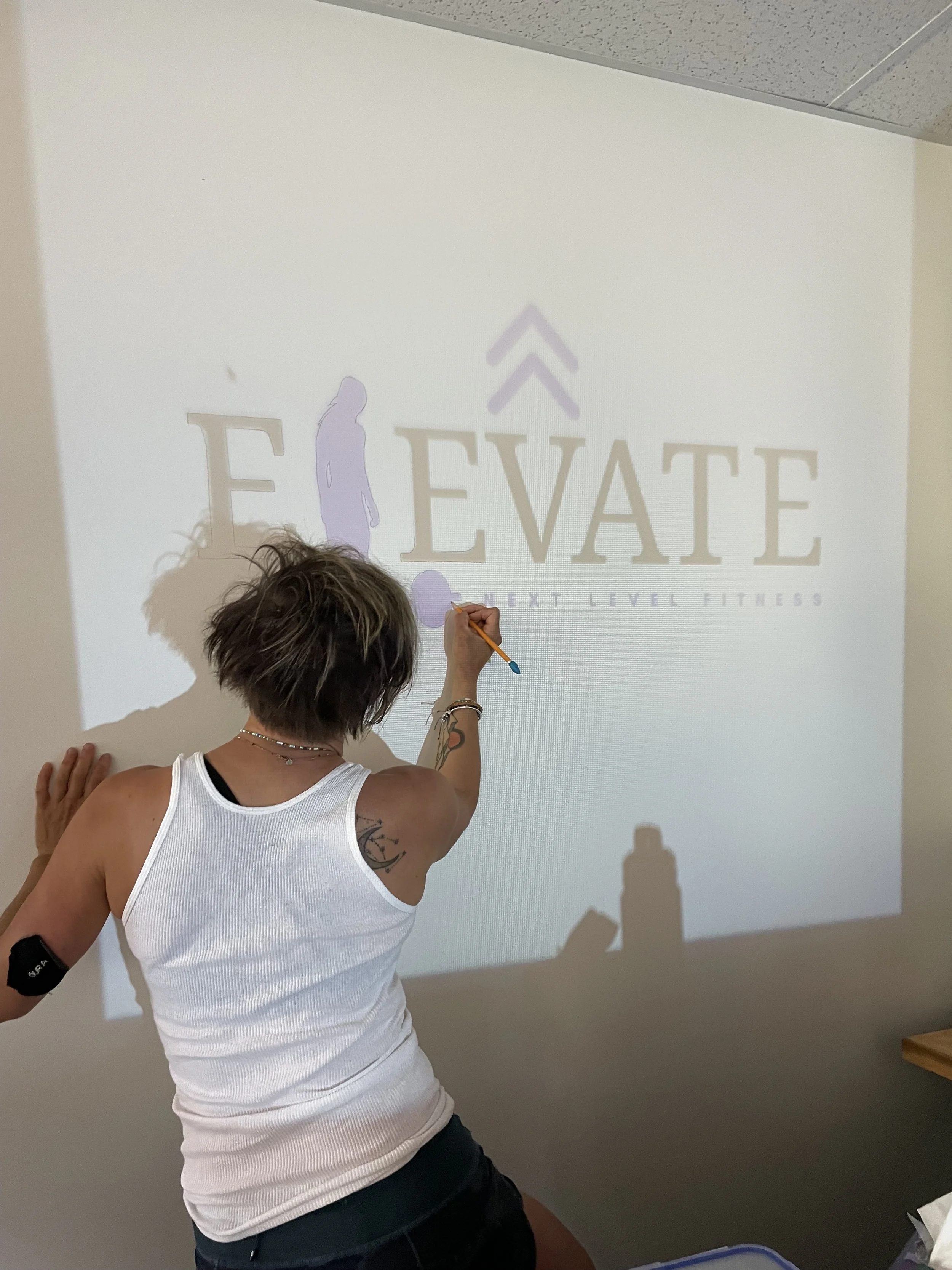 A woman with short hair and tattoos on her arm is drawing on a wall with a marker in front of a projected logo for 'ELEVATE' fitness.