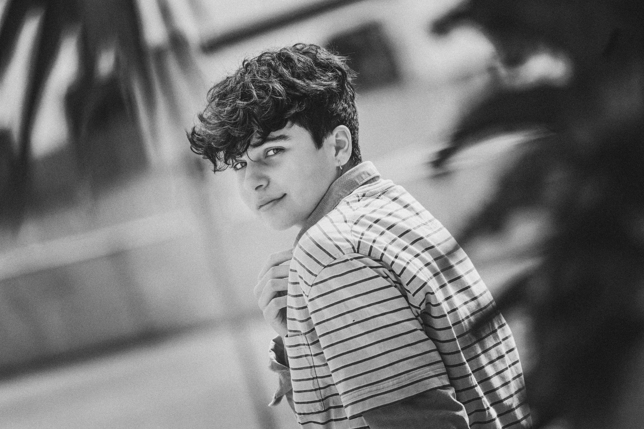 A black and white photo of a young man with curly hair, wearing a striped shirt, looking over his shoulder with a slight smile.
