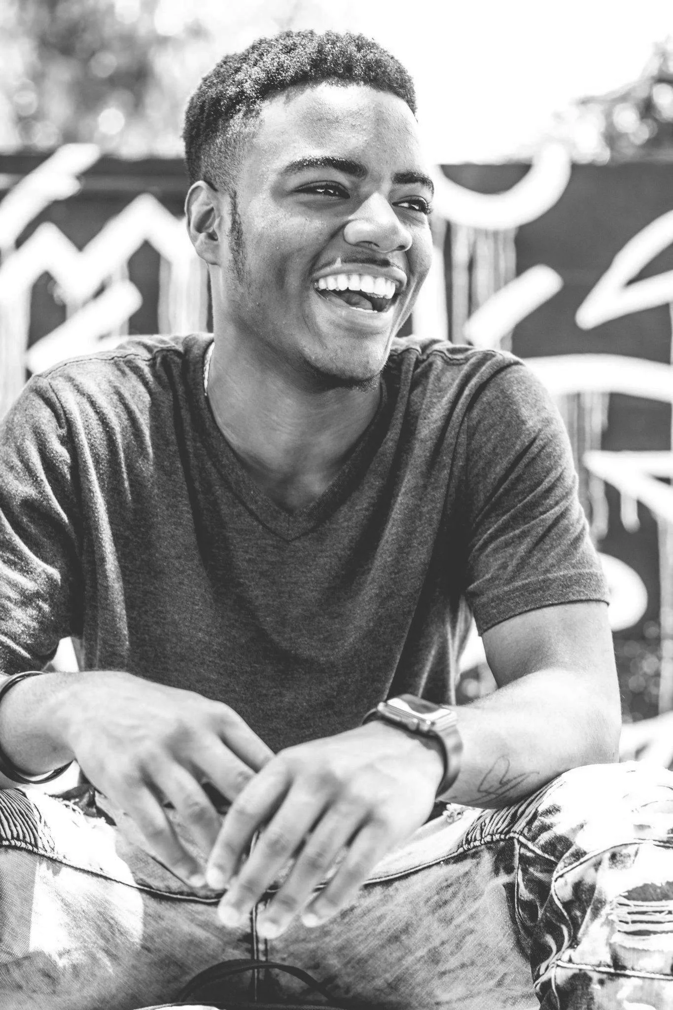 A young man with short curly hair, smiling and laughing while sitting outdoors. He is wearing a dark t-shirt and ripped jeans, with a smartwatch on his left wrist. The background features some abstract graffiti art.